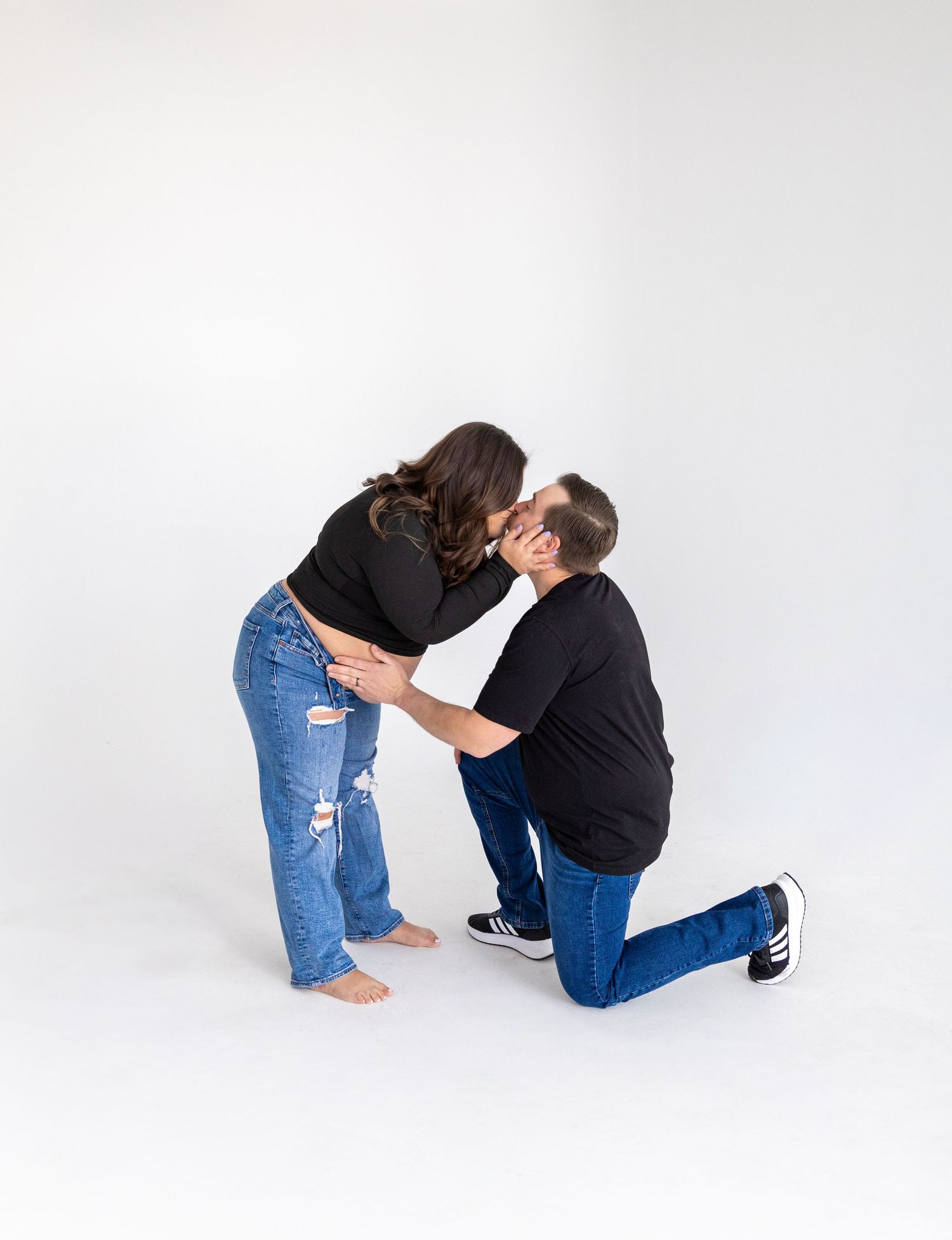 Couple sharing a kiss as man kneels on one knee in a white studio photo session.
