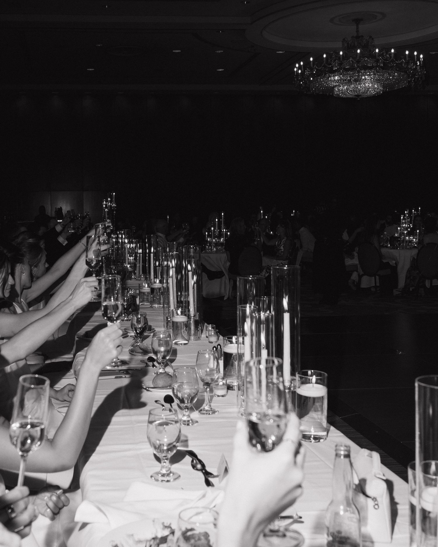 Guests raise champagne glasses in toast along candlelit wedding reception table under crystal chandelier in black and white.