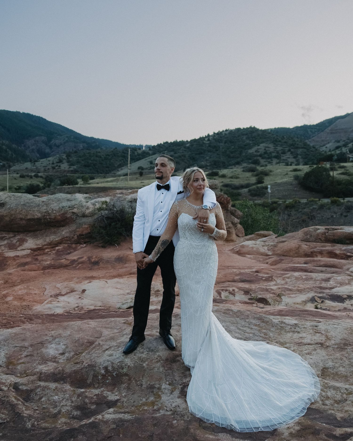 Bride and groom pose on red rocks with mountain backdrop during outdoor Colorado wedding ceremony at dusk.