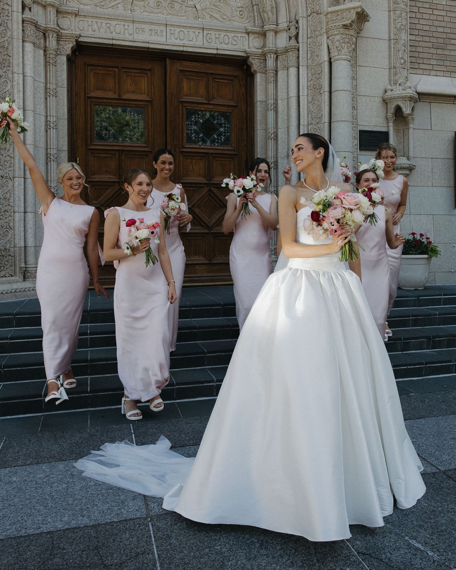 A bride in a white ball gown laughs as bridesmaids in pink dresses celebrate on church steps.