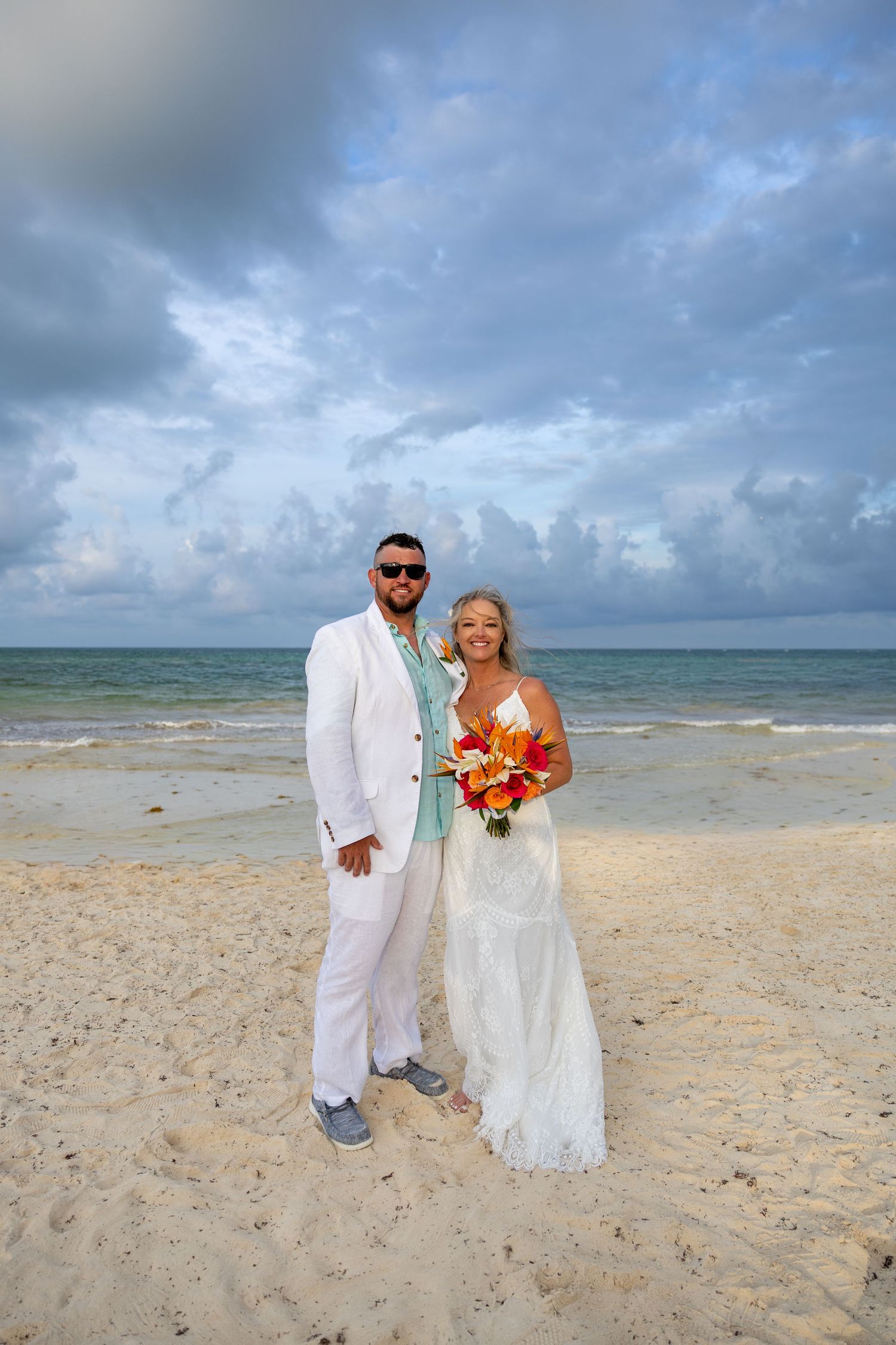 Newlyweds in white wedding attire standing on beach with cloudy sky backdrop.