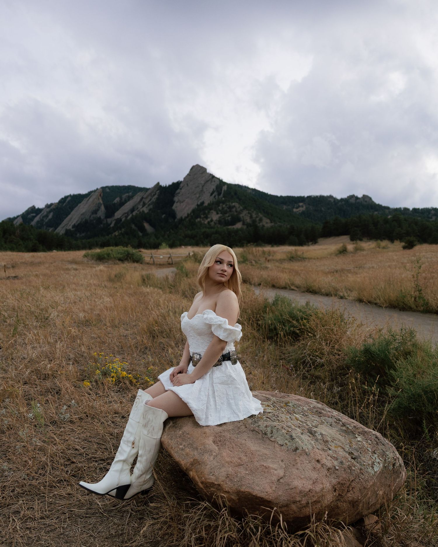 Bride in white mini dress and cowboy boots sits on boulder with Flatirons mountain backdrop in Colorado.