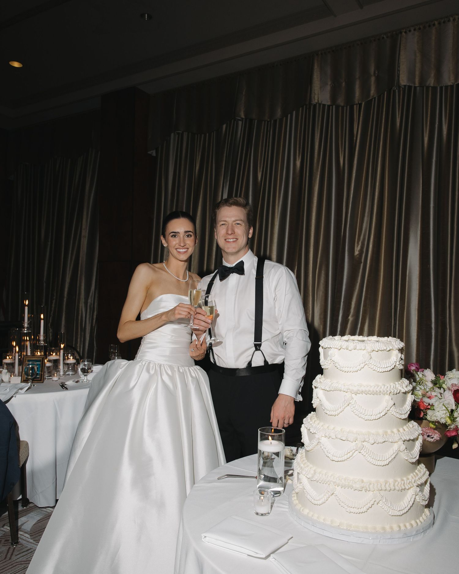 Bride and groom smiling beside four-tier white wedding cake at elegant indoor reception venue.
