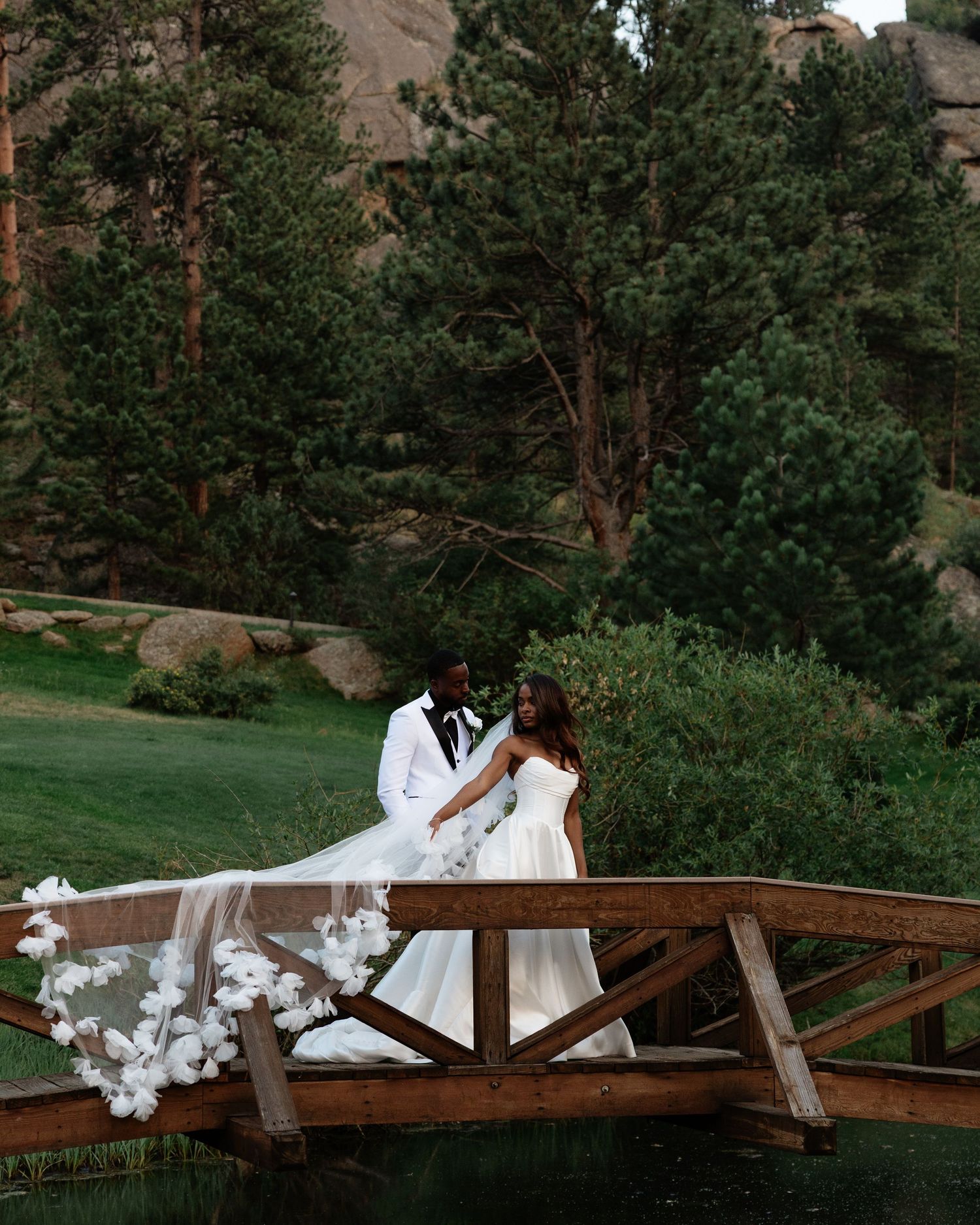 Bride and groom walking on a wooden bridge with a dramatic floral veil and mountains.