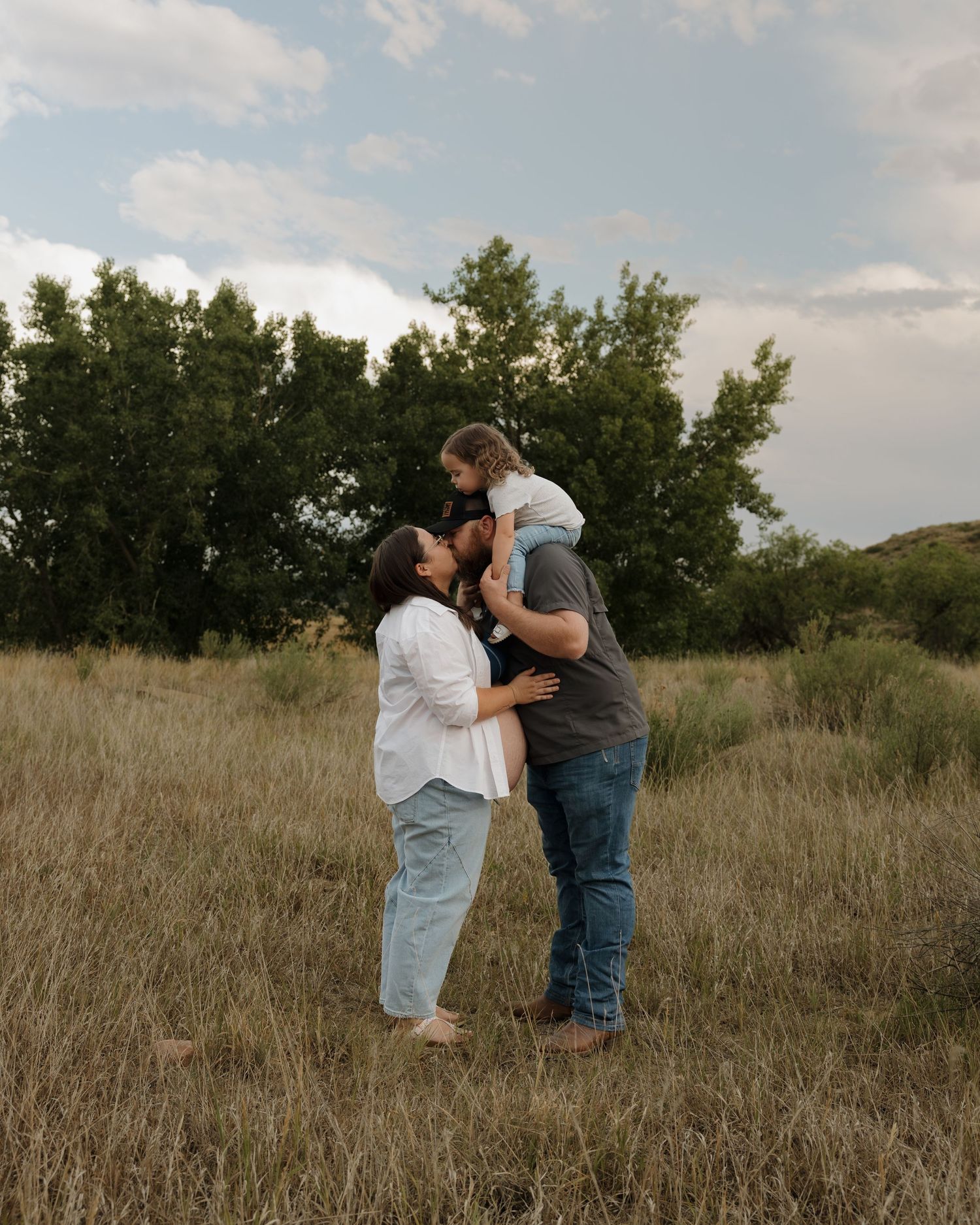 A family of three poses in a golden meadow, parents kissing as dad lifts toddler overhead against a cloudy sky. Beautiful family photography