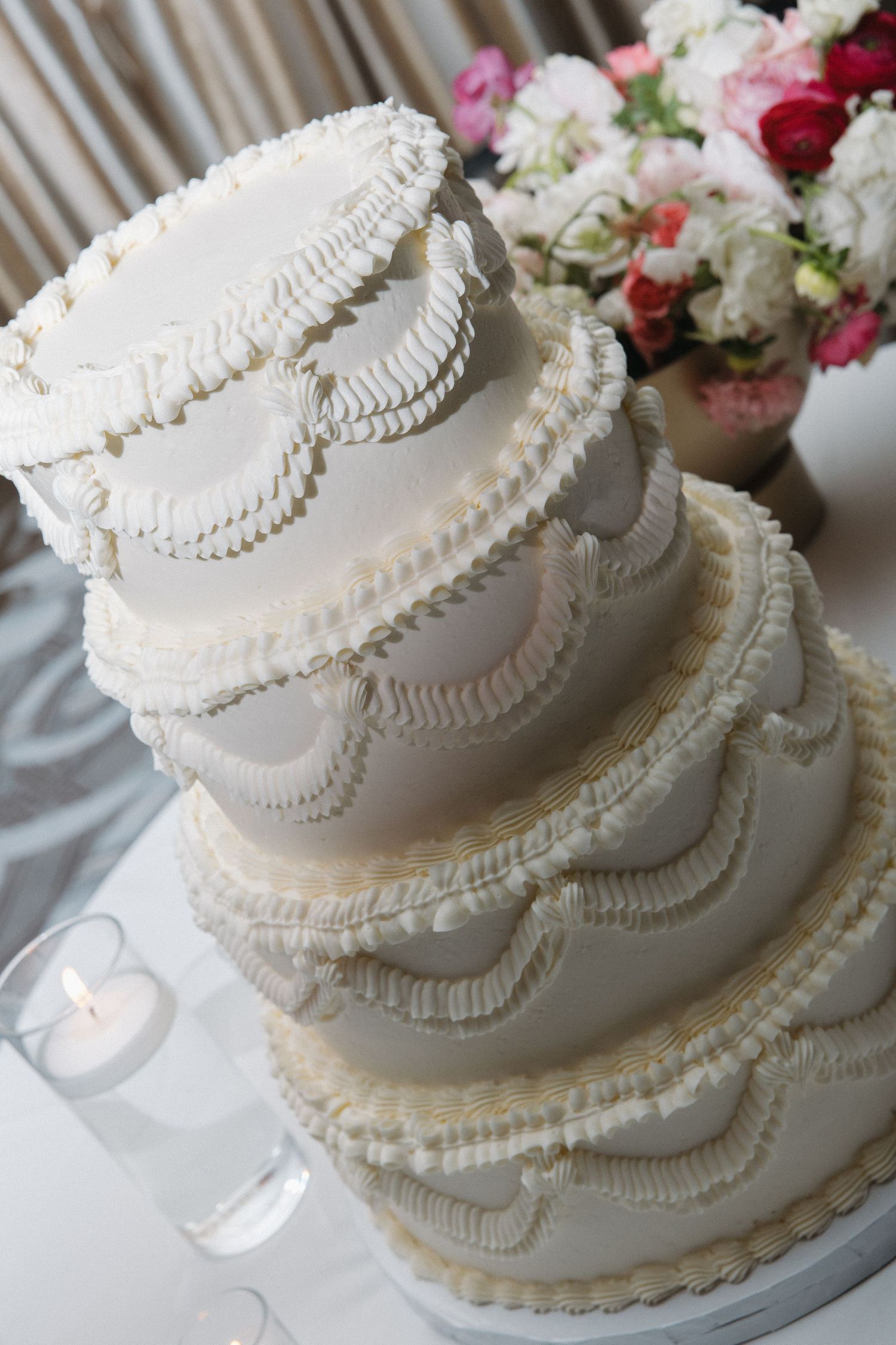 Close-up of elegant four-tier white wedding cake with ruffled frosting and floral arrangement nearby.