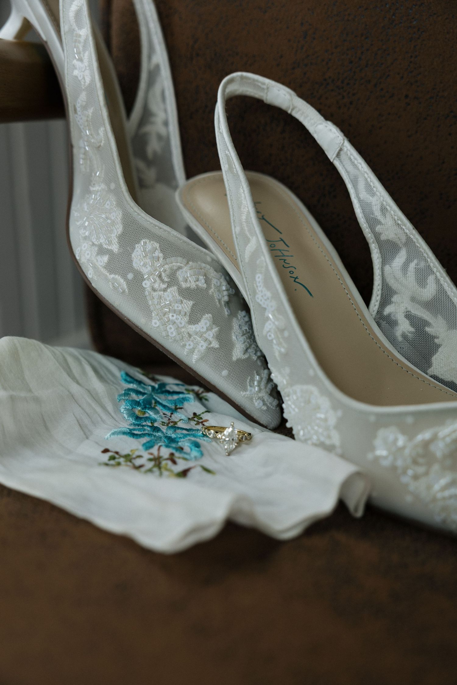 Elegant white lace bridal heels displayed with an embroidered handkerchief and gold ring on a brown chair.