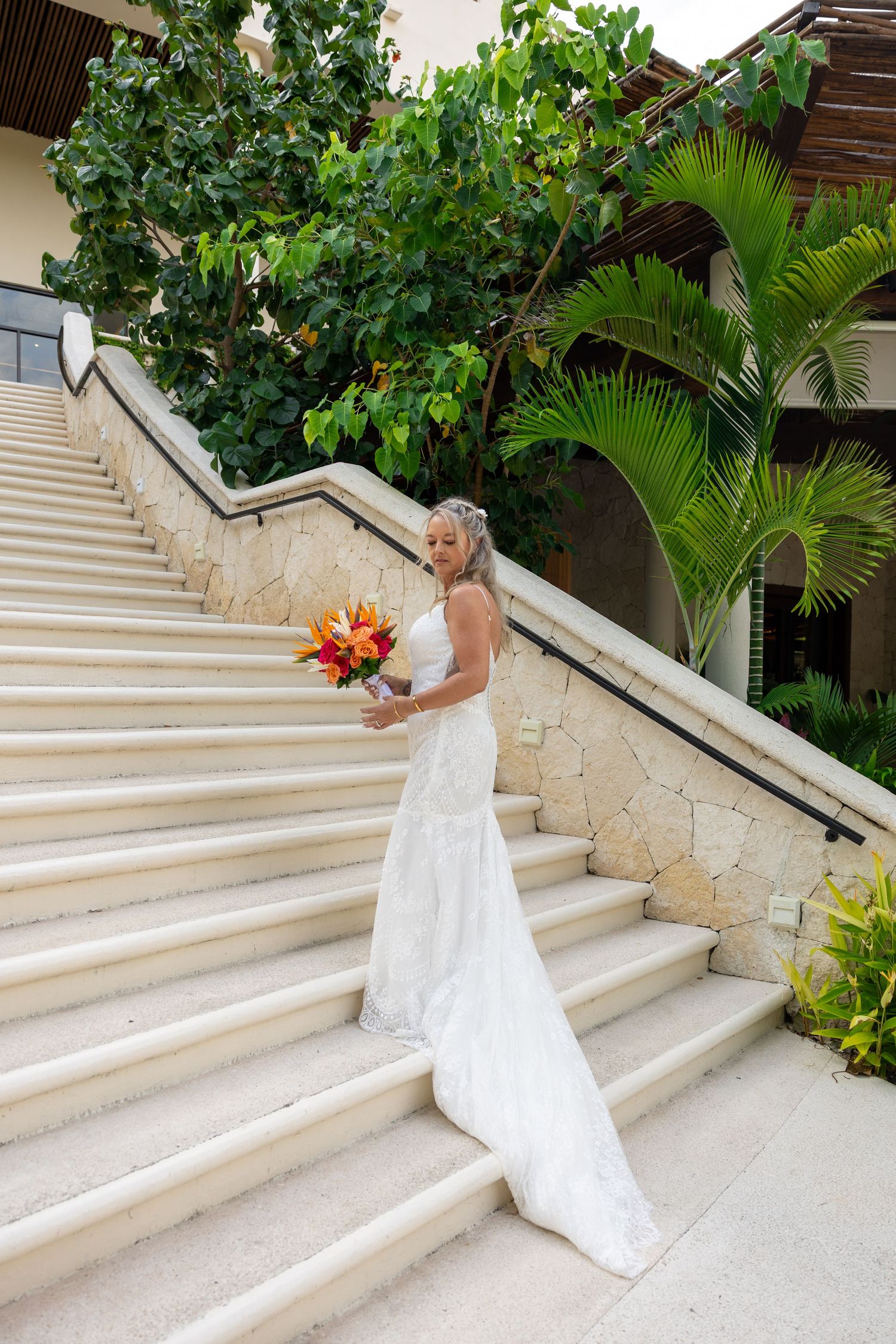 Elegant bride in lace gown holding tropical bouquet on grand stone staircase.