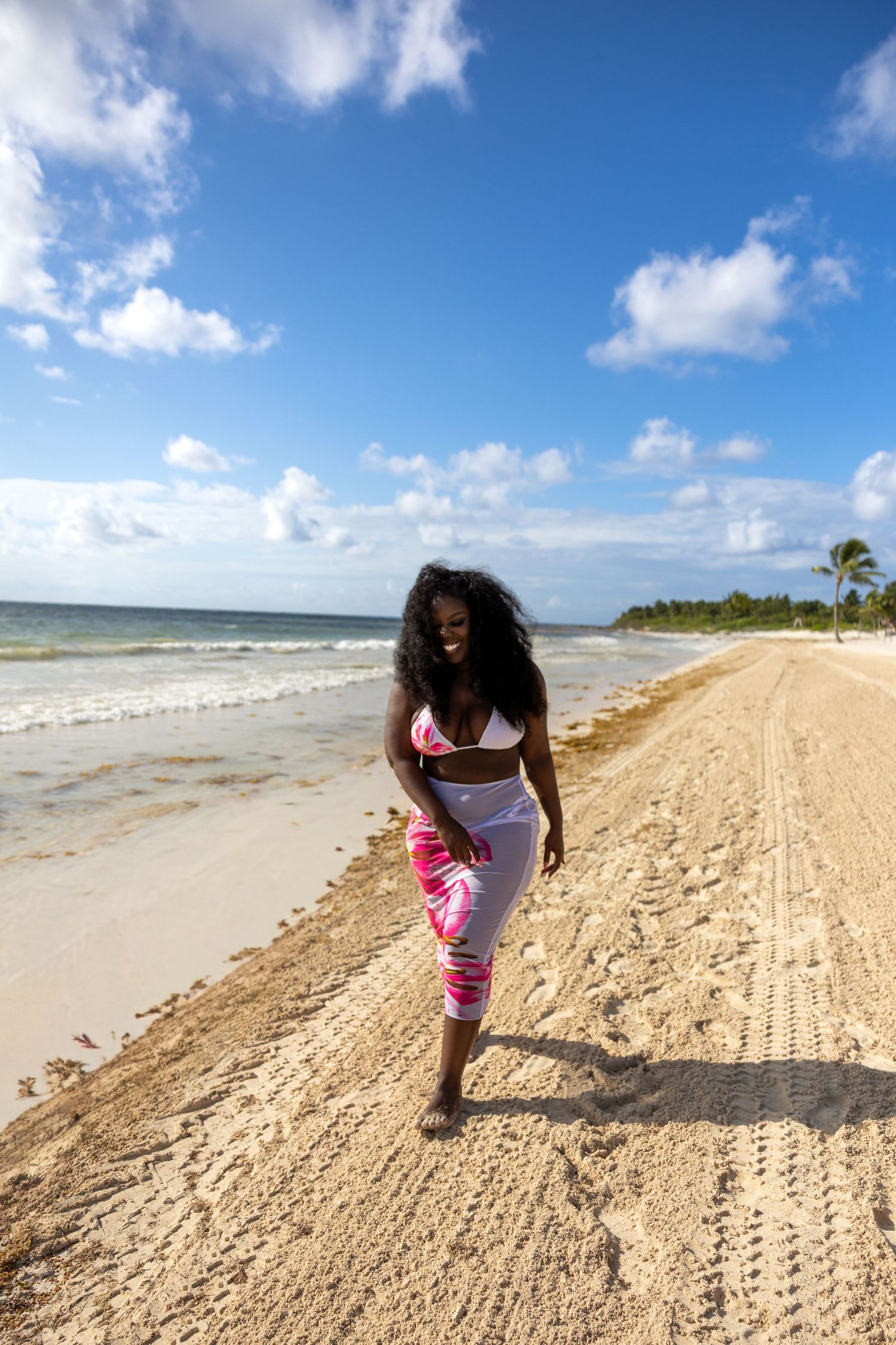 Black woman in pink bikini top and tie-dye pants walking barefoot along sunny tropical beach shoreline.