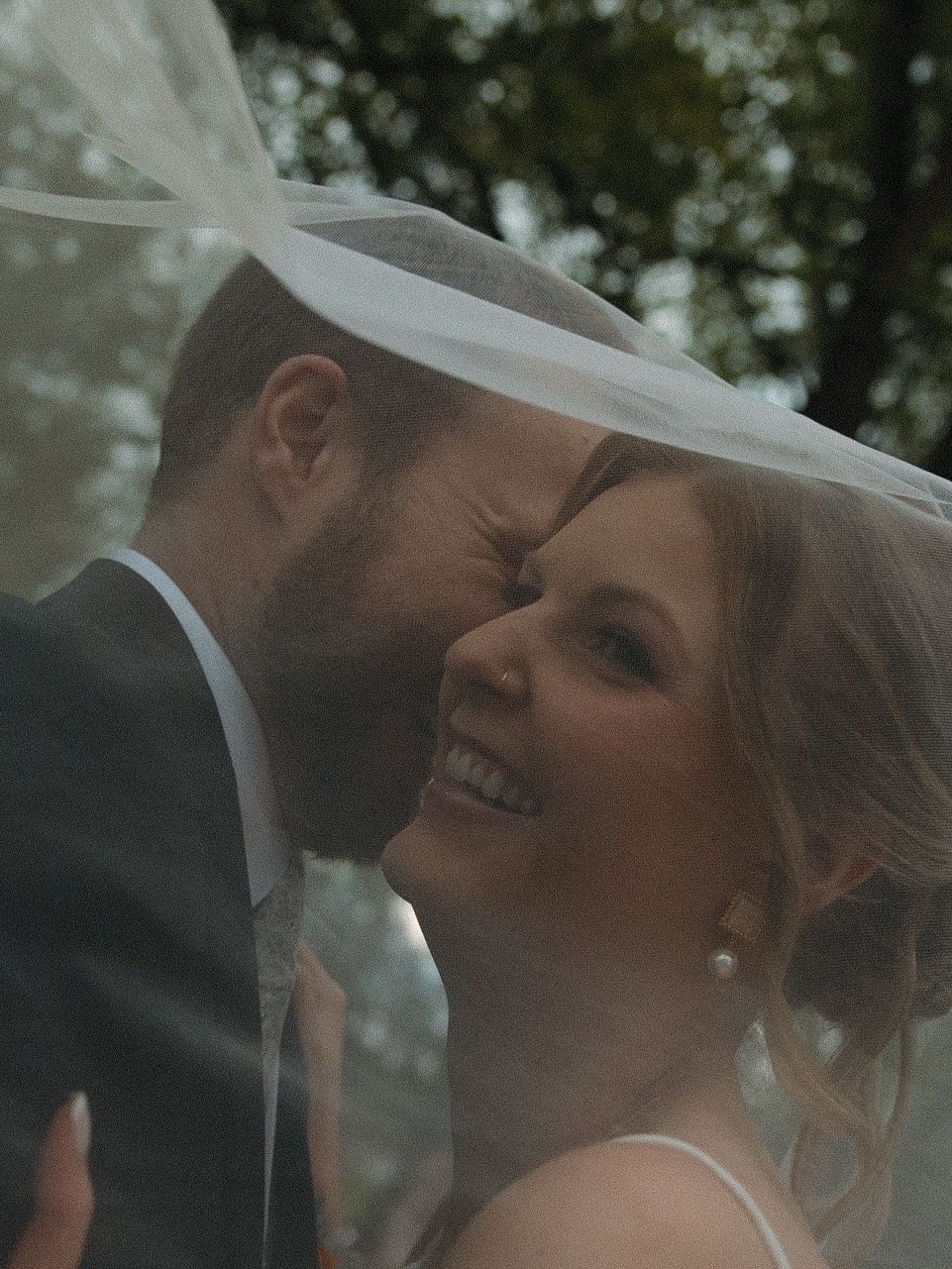A groom kisses a laughing bride on the cheek under a flowing veil in a romantic outdoor portrait.
