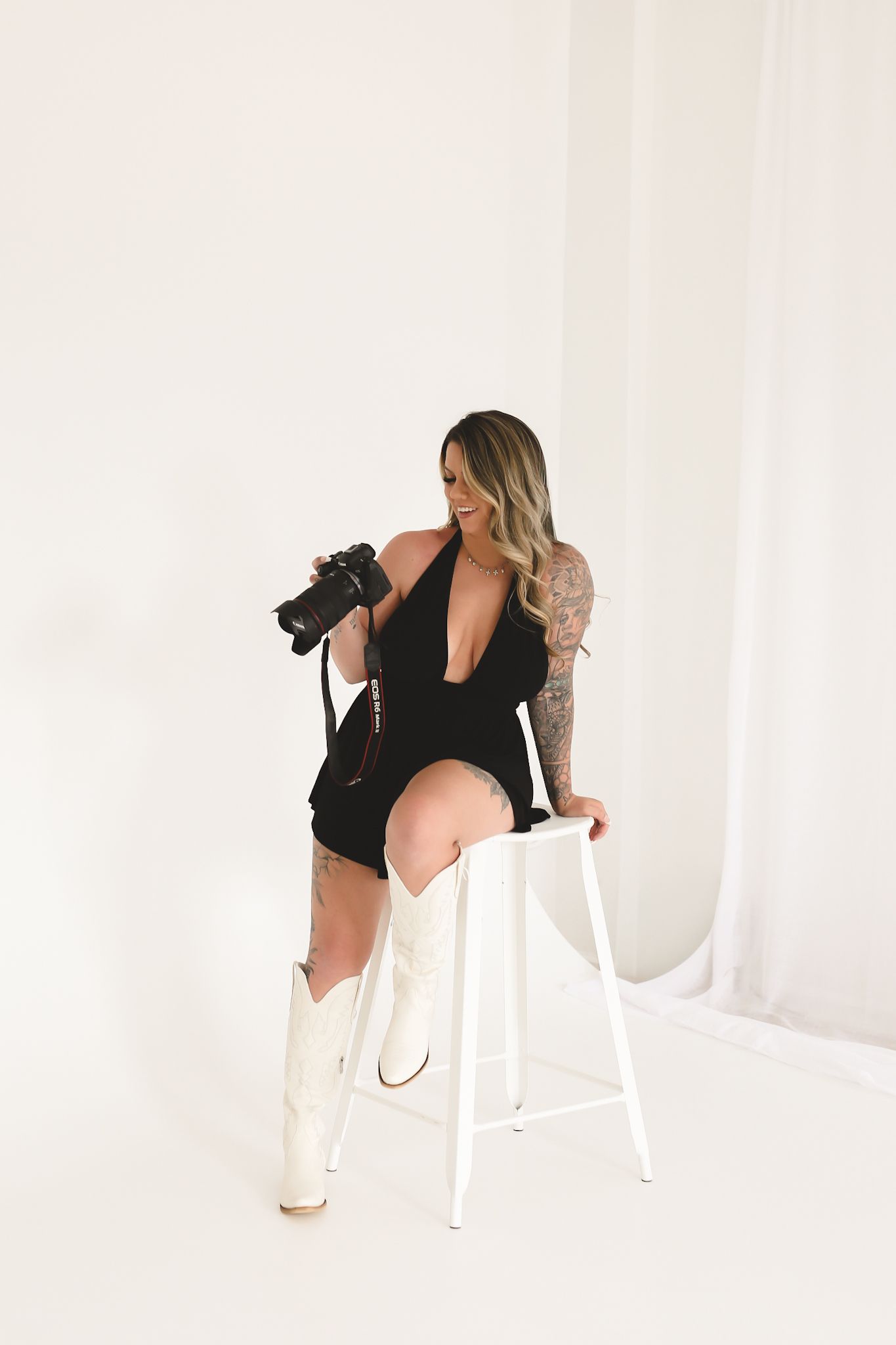 Smiling tattooed photographer in black romper and white cowboy boots reviews Canon camera on stool.