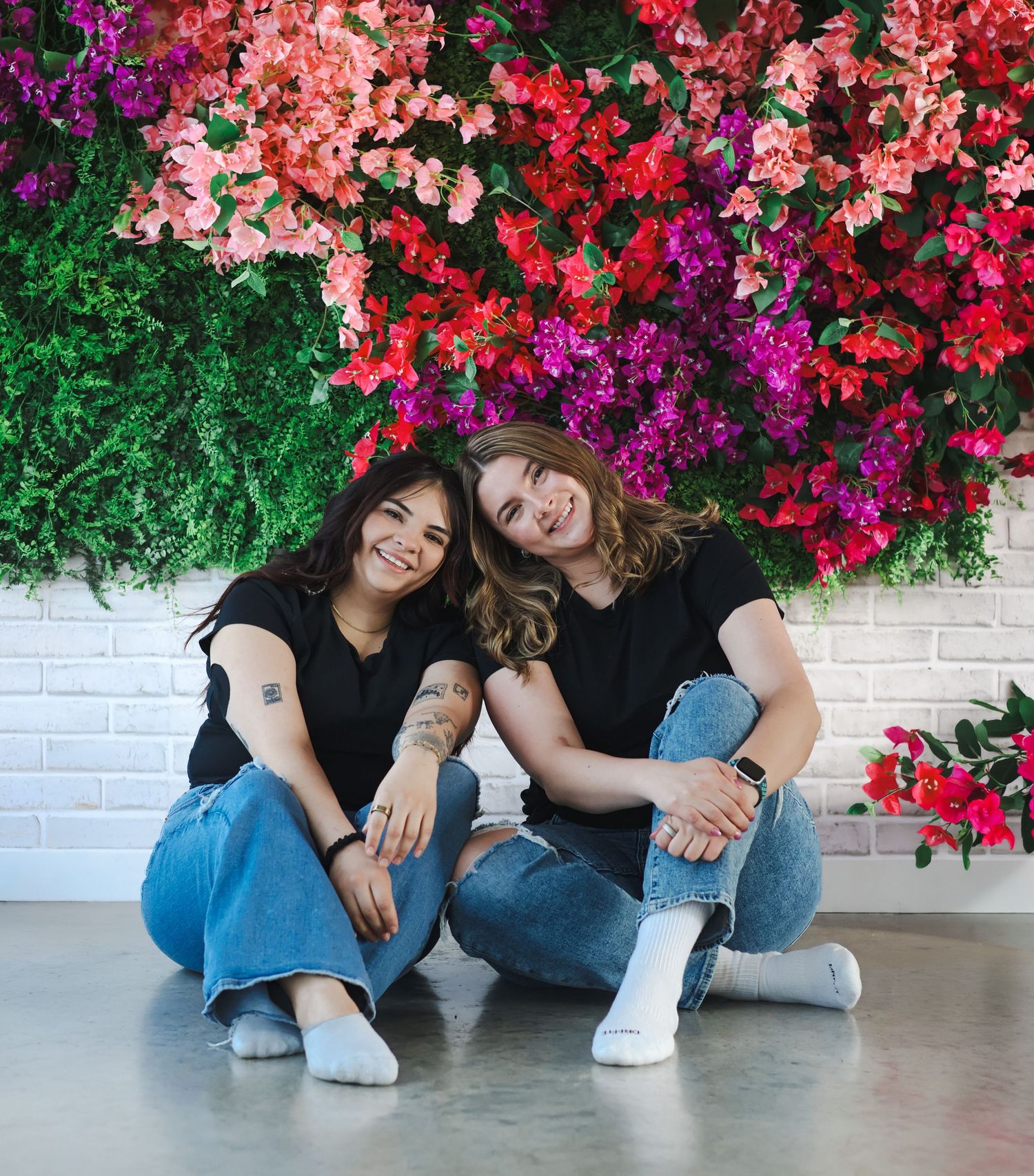 Two women sitting together smiling in front of vibrant flower wall backdrop.