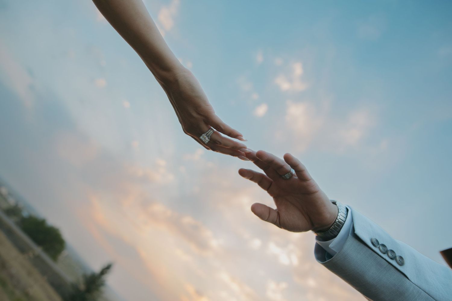 Bride and groom reaching toward each other against a pastel sunset sky.