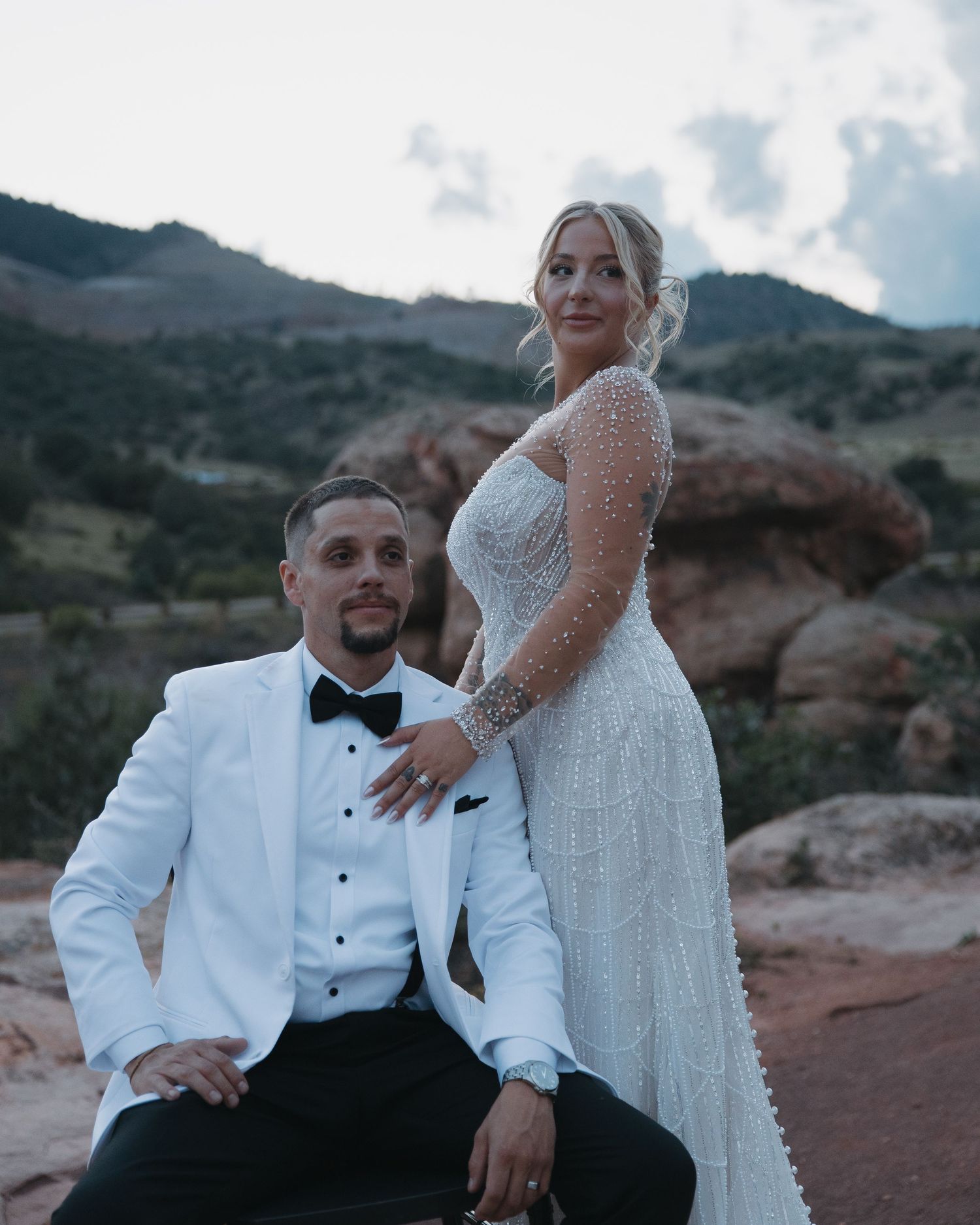 Wedding couple posing at red rocks, groom seated in white tuxedo, bride standing in beaded gown.