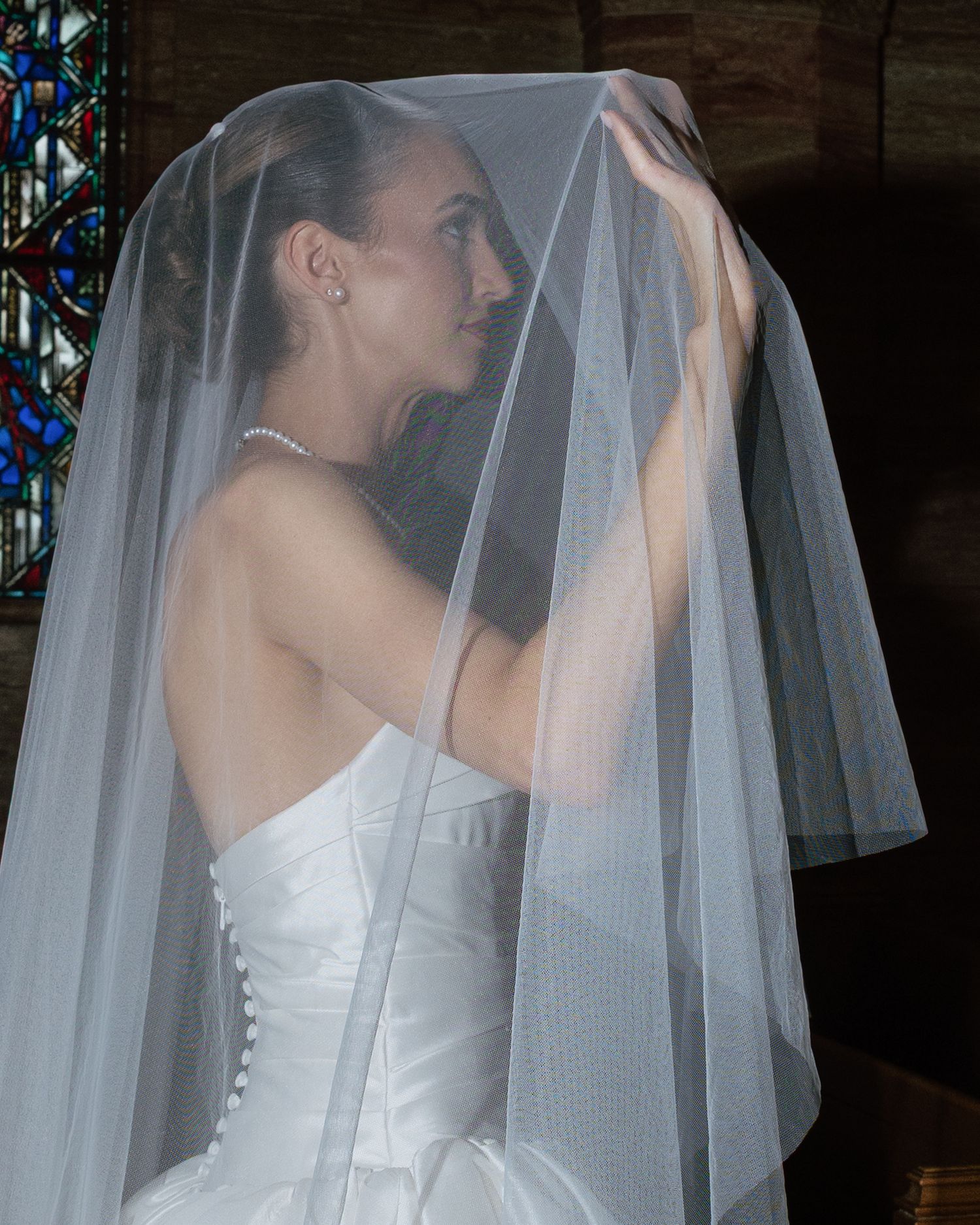 Bride in strapless white gown lifts sheer veil inside church with stained glass window behind her.