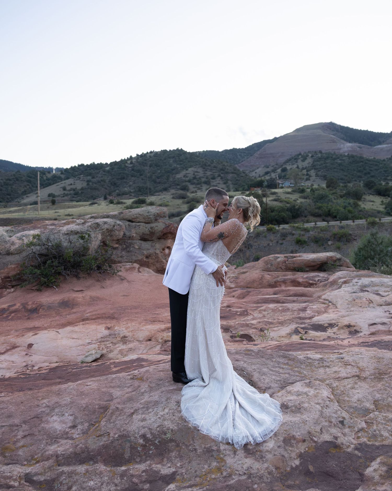 Bride and groom sharing romantic kiss on red rock formation with mountain landscape at dusk.