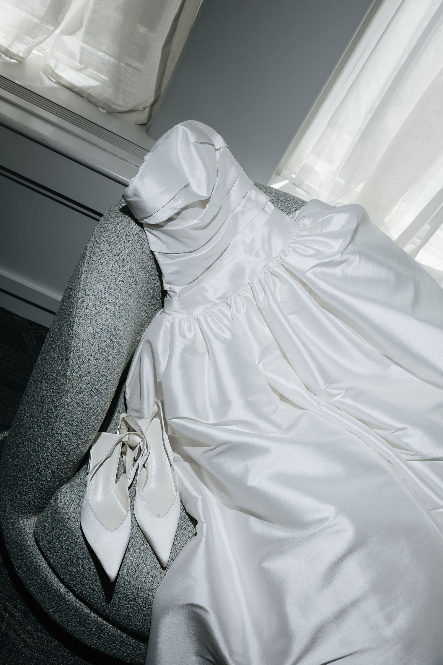 White wedding gown and heels draped over gray chair by window.
