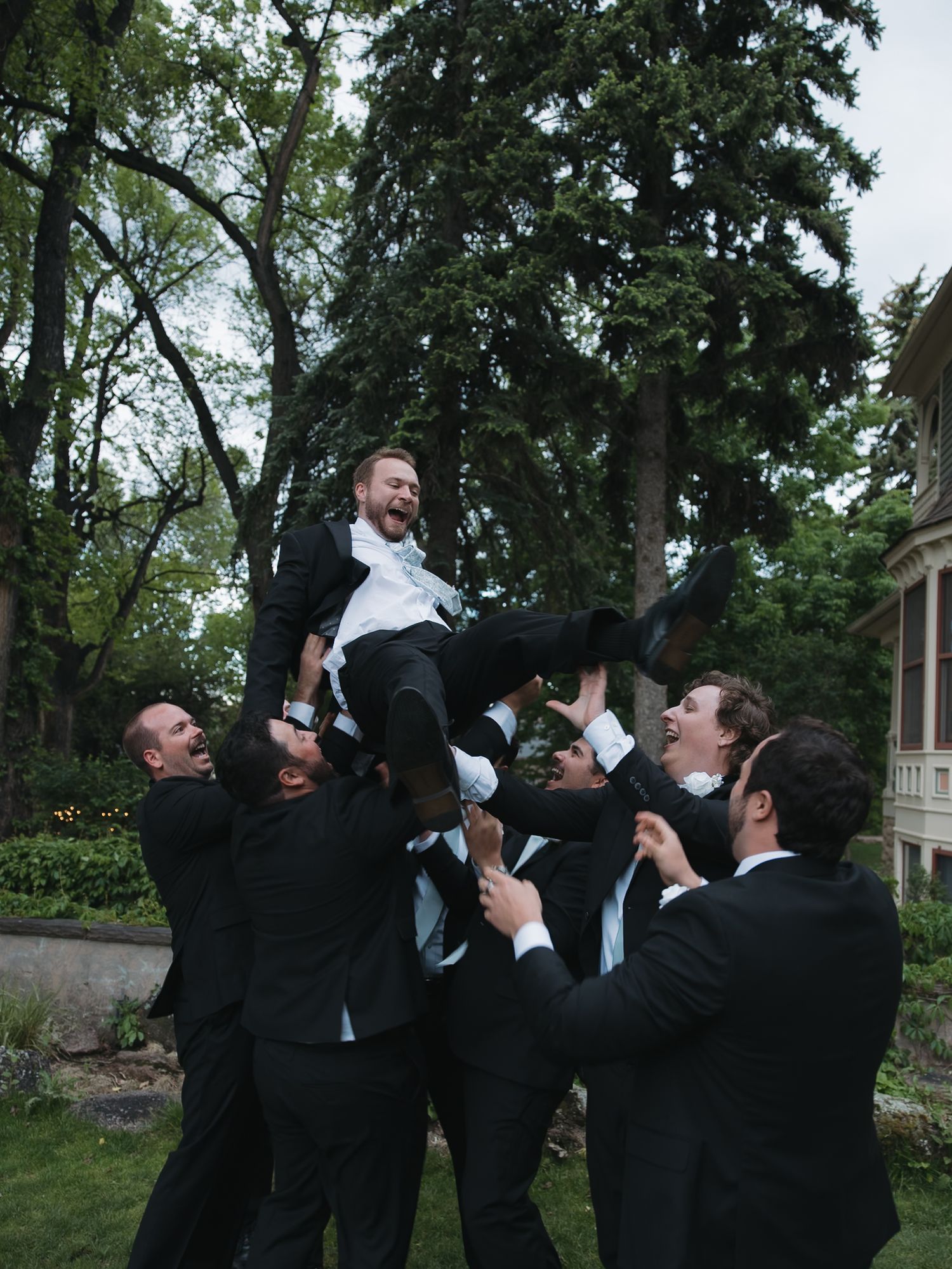 Groomsmen toss groom into air outdoors in celebratory wedding photo.