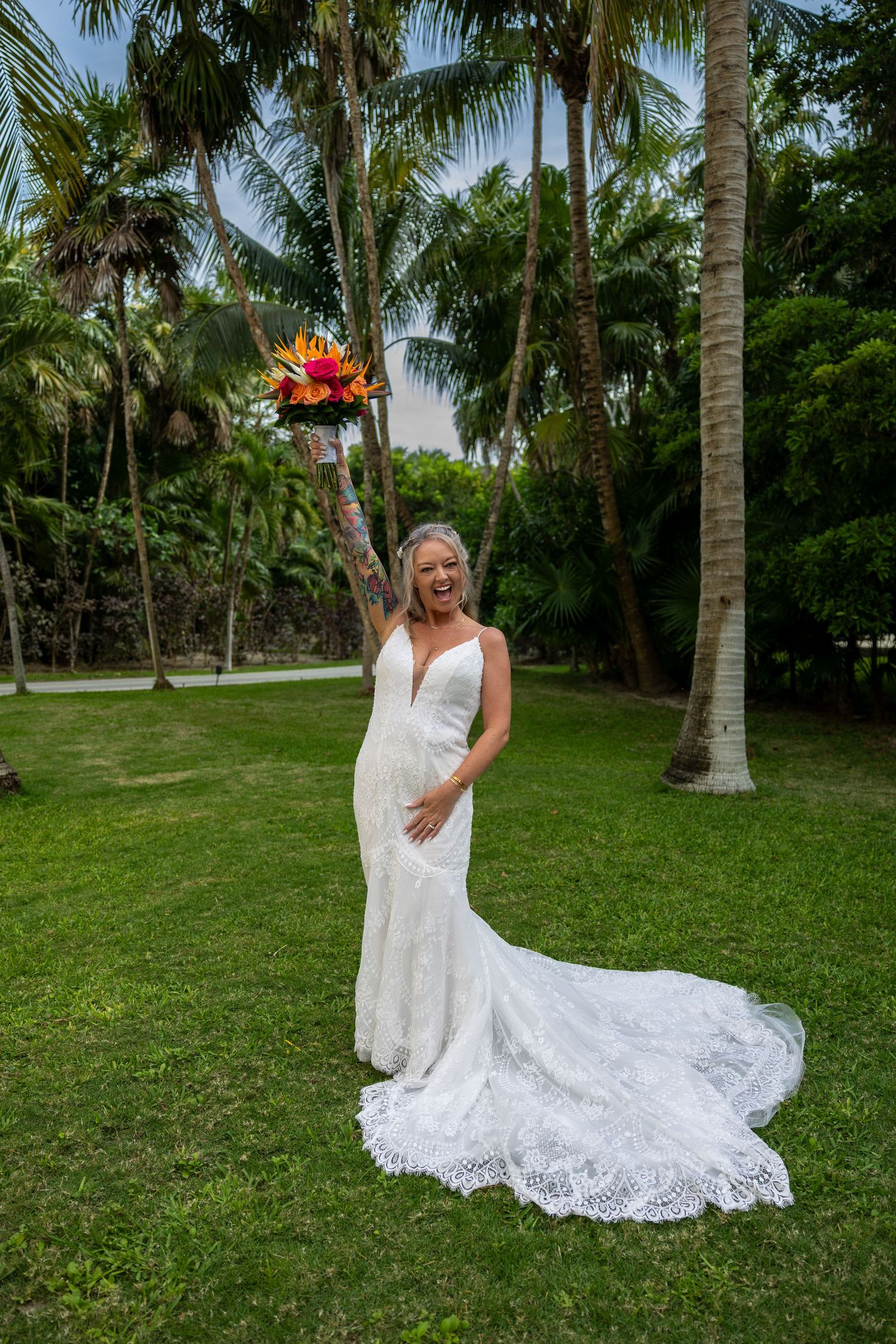 Joyful bride in white lace gown raises colorful bouquet among palm trees on lush green lawn.