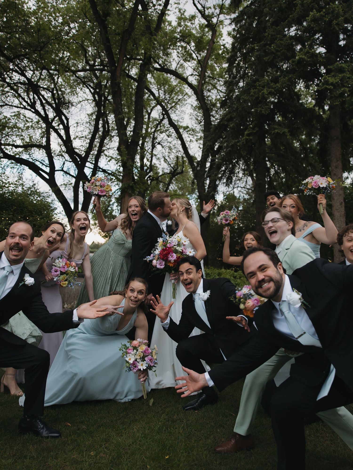 Wedding party celebrating outdoors, bride and groom kissing surrounded by bridesmaids in mint dresses and groomsmen in black suits.