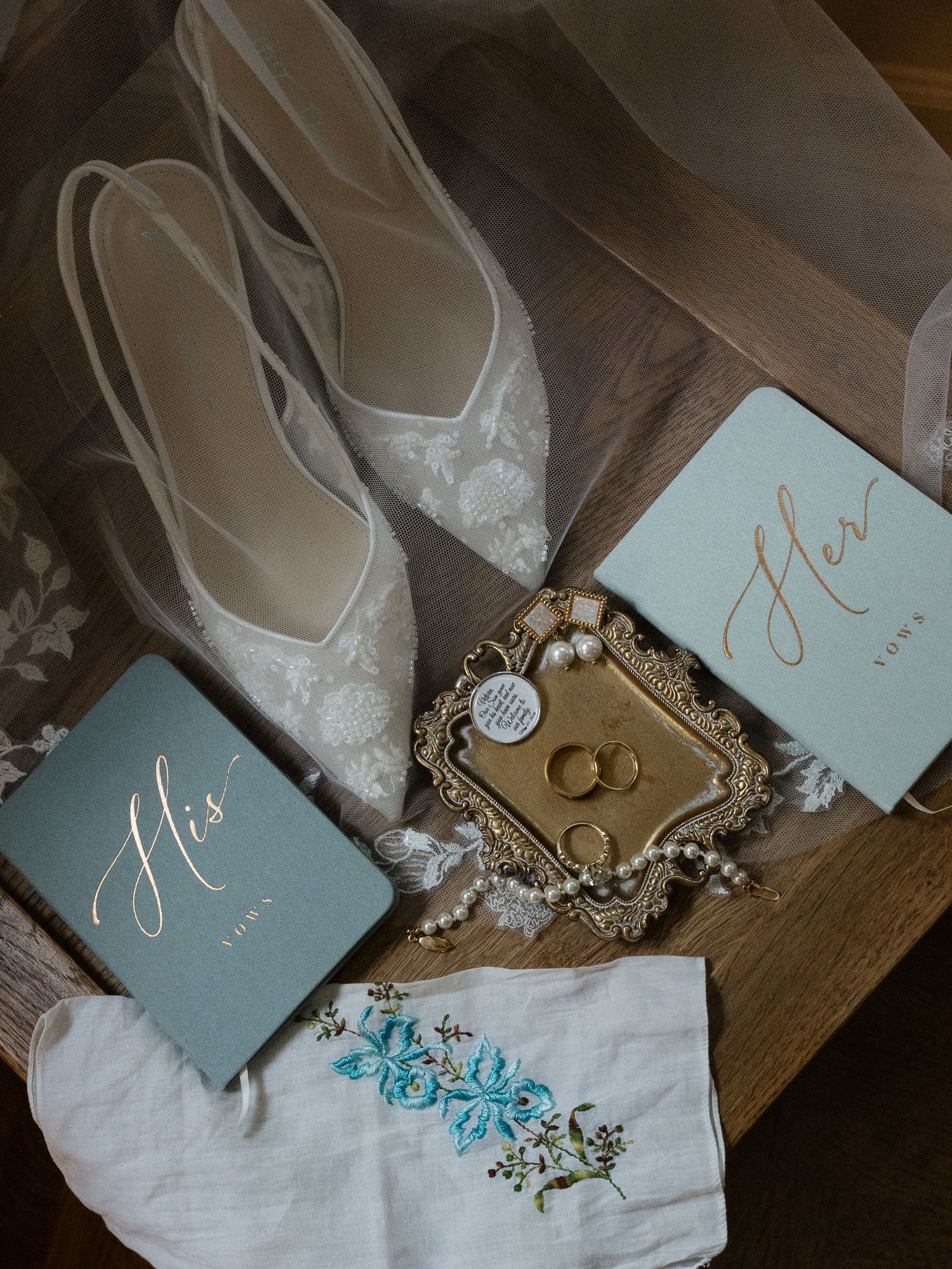Elegant wedding flat lay with lace heels, gold ring dish, pearl bracelet, His and Hers vow books, and embroidered handkerchief.