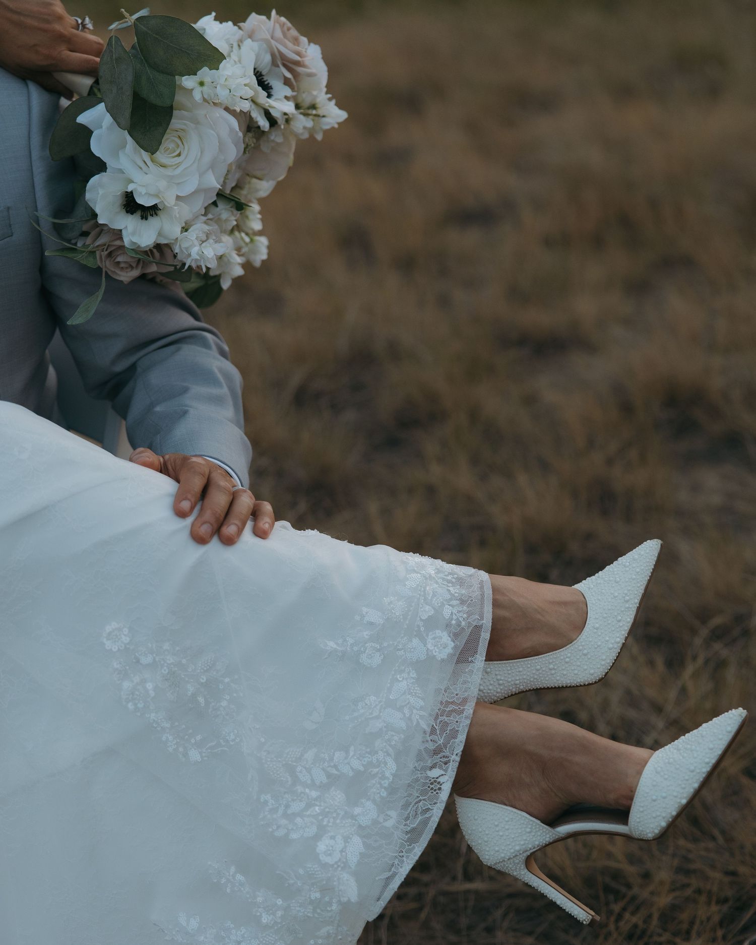 Bride in white lace gown and heels sits with groom holding a blush bouquet in a field.