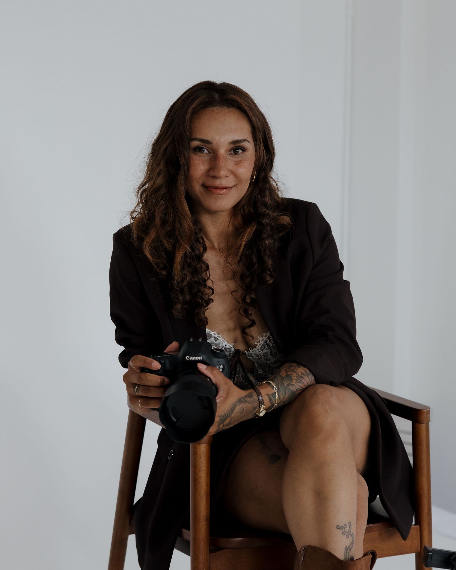 Female wedding photographer with curly hair sits on wooden chair holding Canon camera in studio. Branding sessions