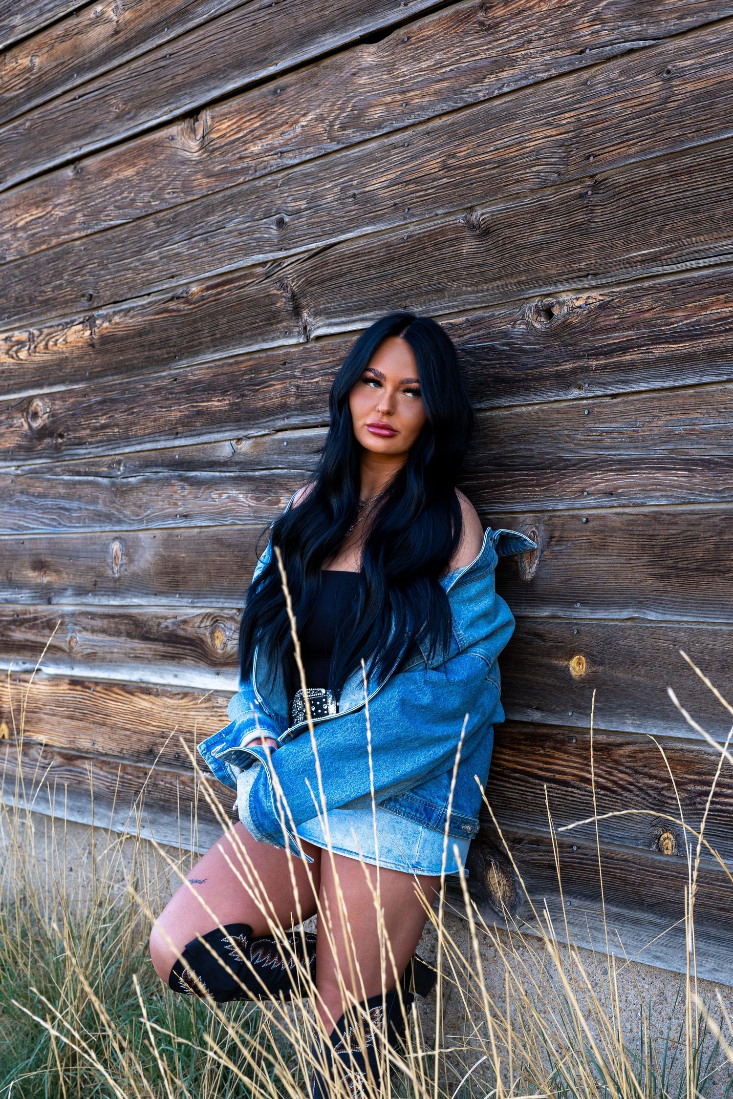 Young woman with long black hair in denim jacket and mini skirt leaning against rustic wooden barn wall.