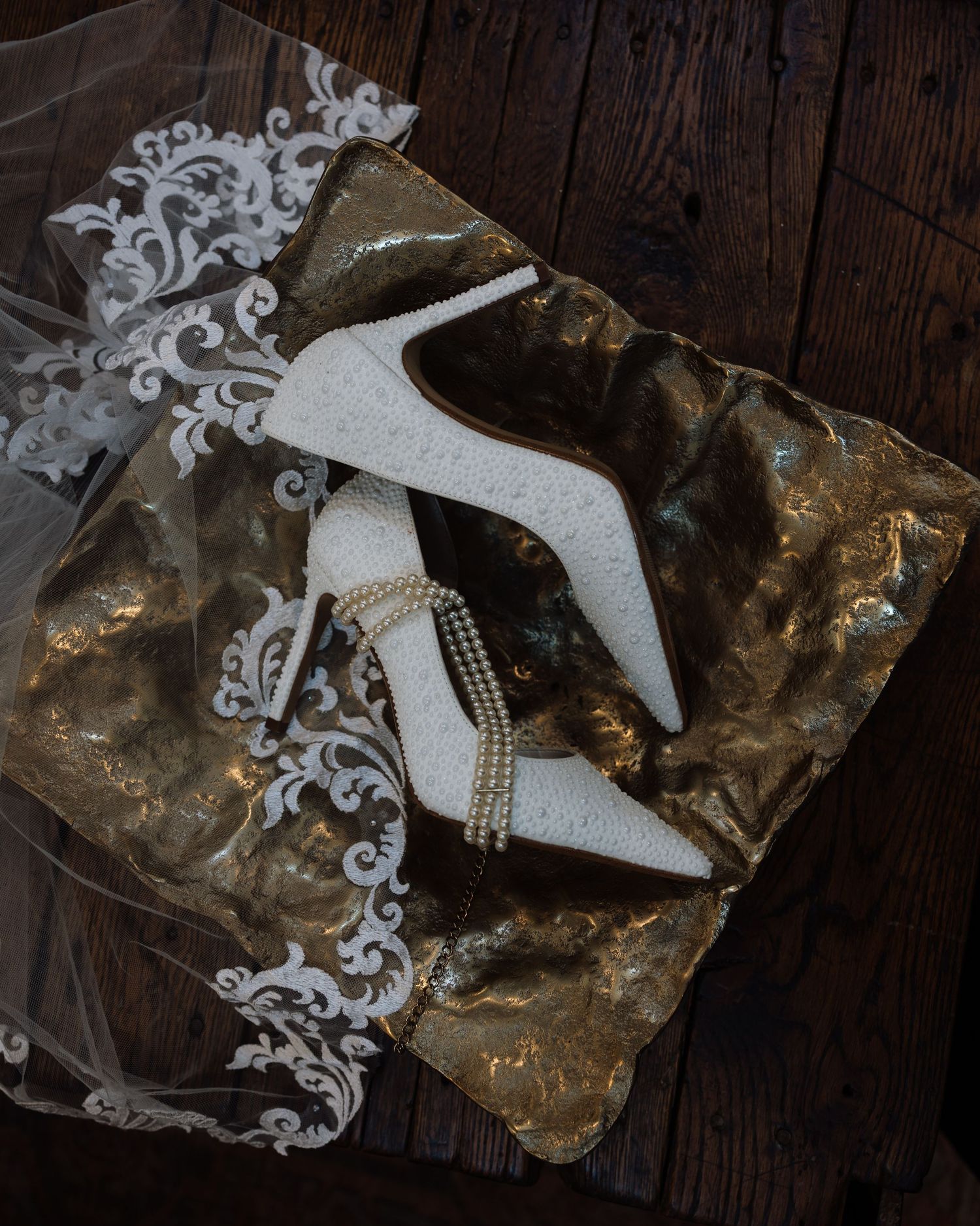 White lace bridal heels with pearl ankle straps displayed on gold tissue paper beside a wedding veil.