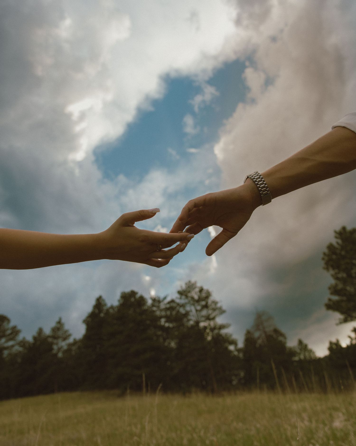 Two hands reaching toward each other against a cloudy sky with green trees below.