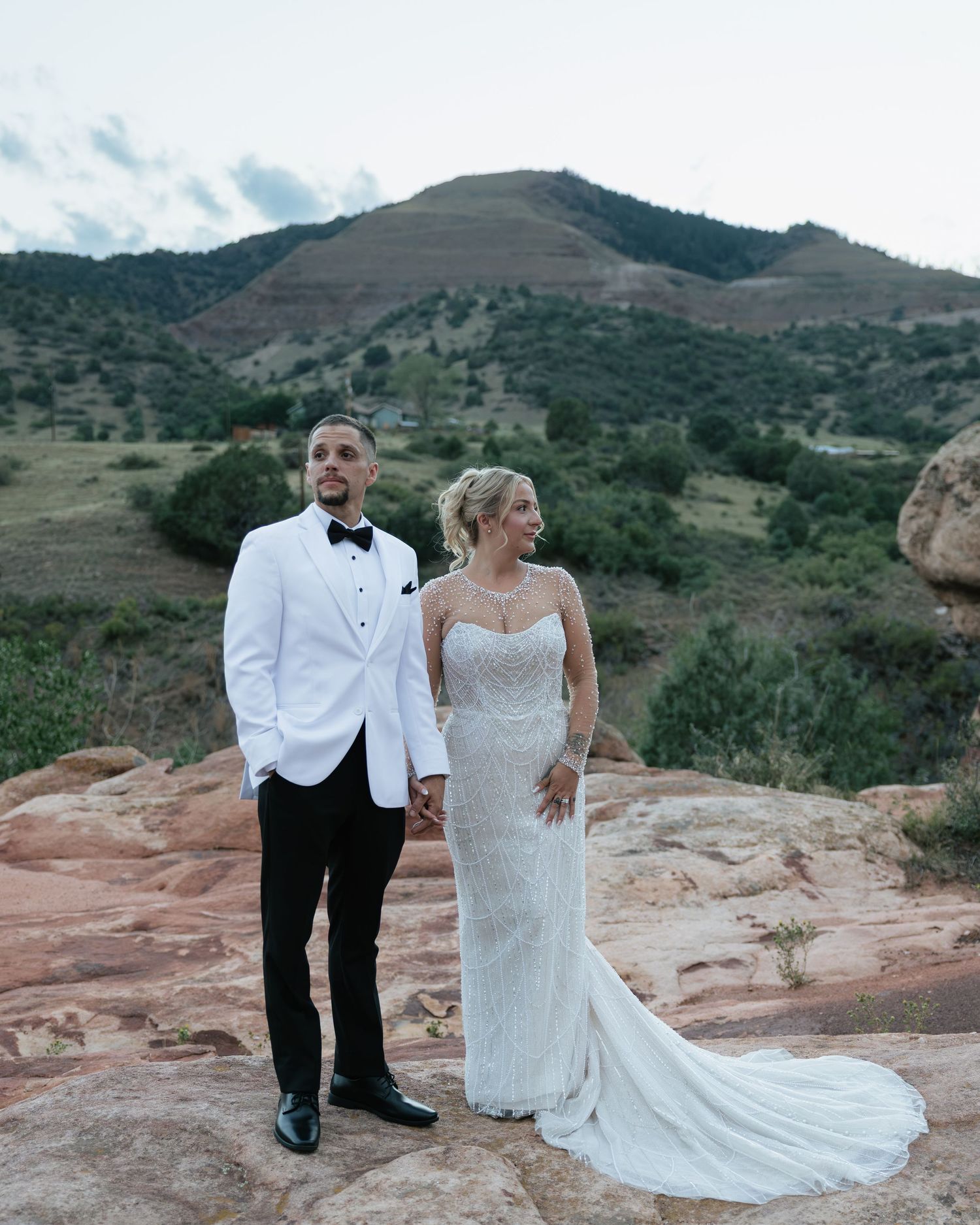 Bride and groom hold hands posing on red rocks with scenic mountain landscape backdrop.