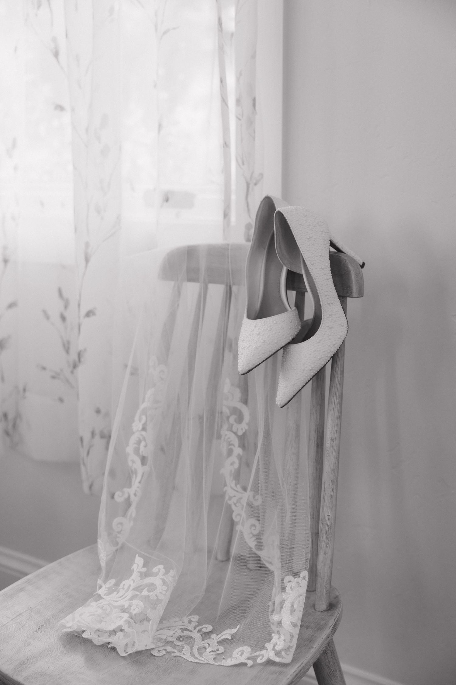 Black and white photo of bridal heels and lace veil draped over wooden chair.