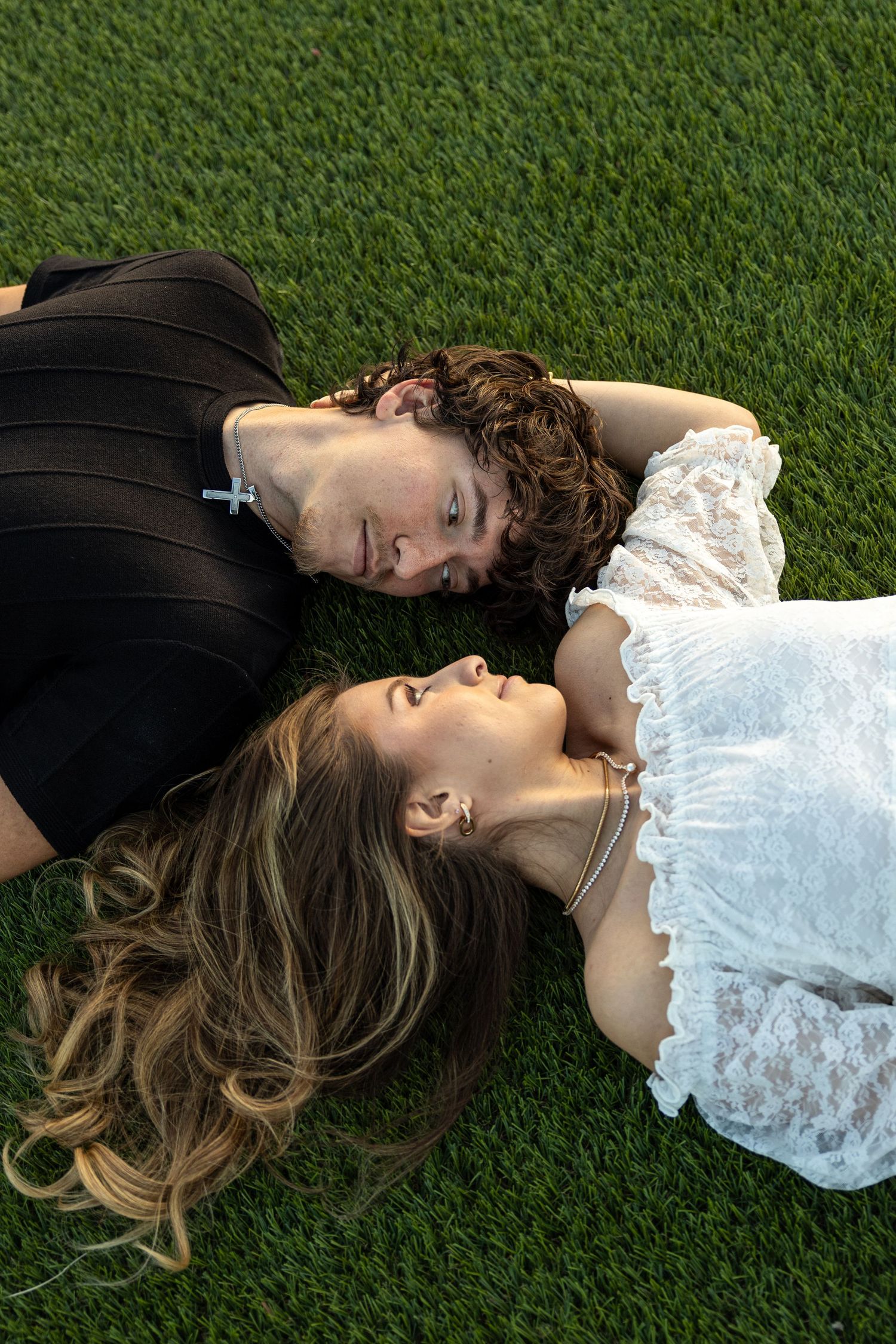 Couple lying on green grass facing each other in a romantic outdoor engagement photo.