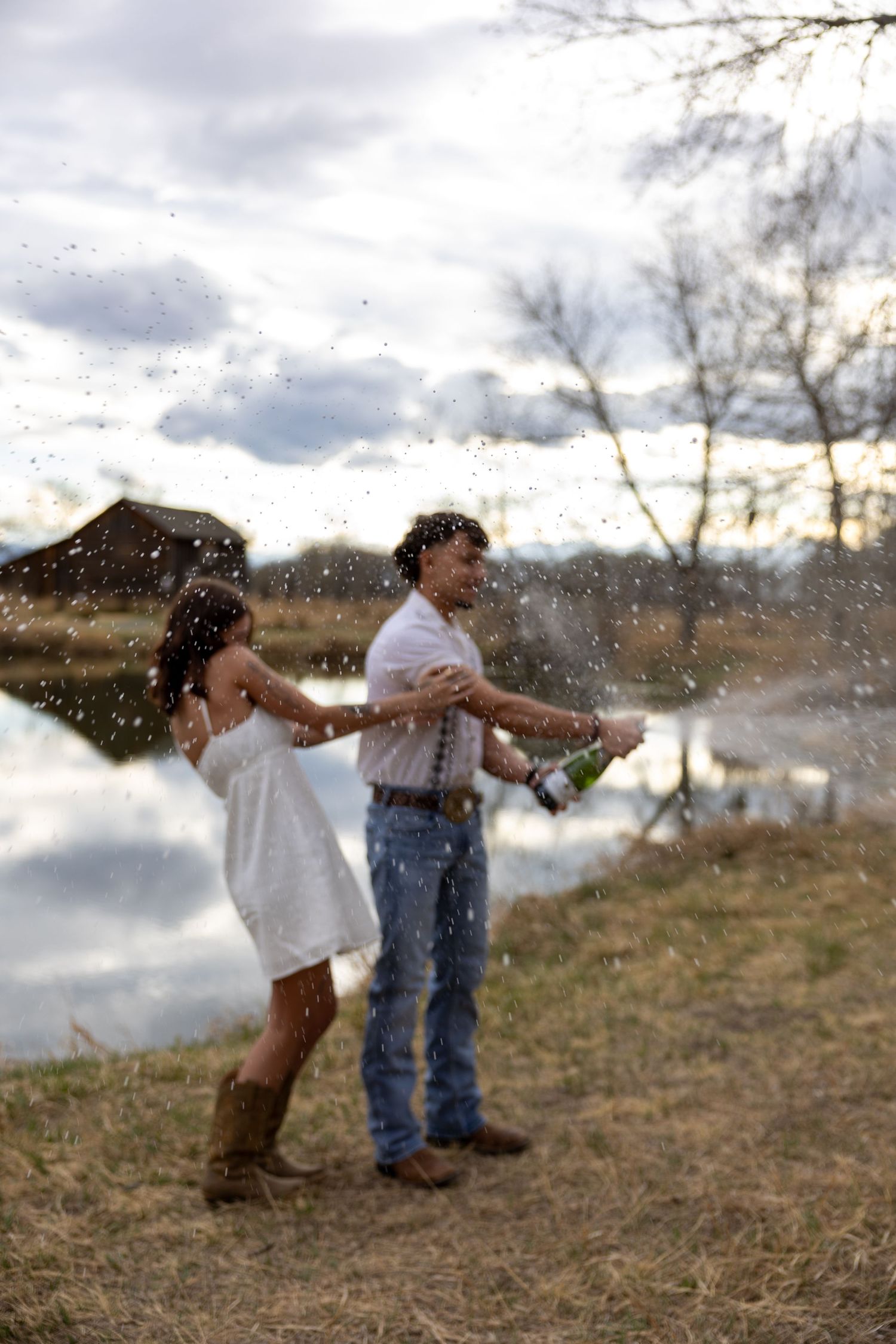 A couple celebrates by a reflective pond, popping champagne as droplets spray around them at dusk. Elegant engagement photography