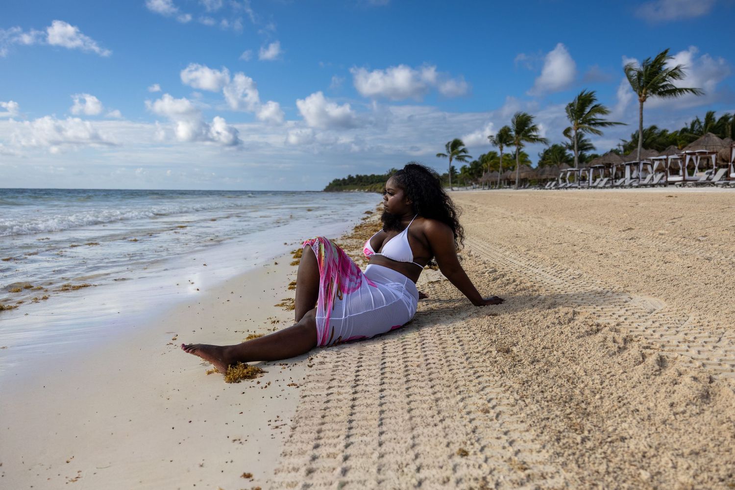 Woman in white bikini top and pink sarong sitting relaxed on a sandy tropical beach shoreline.