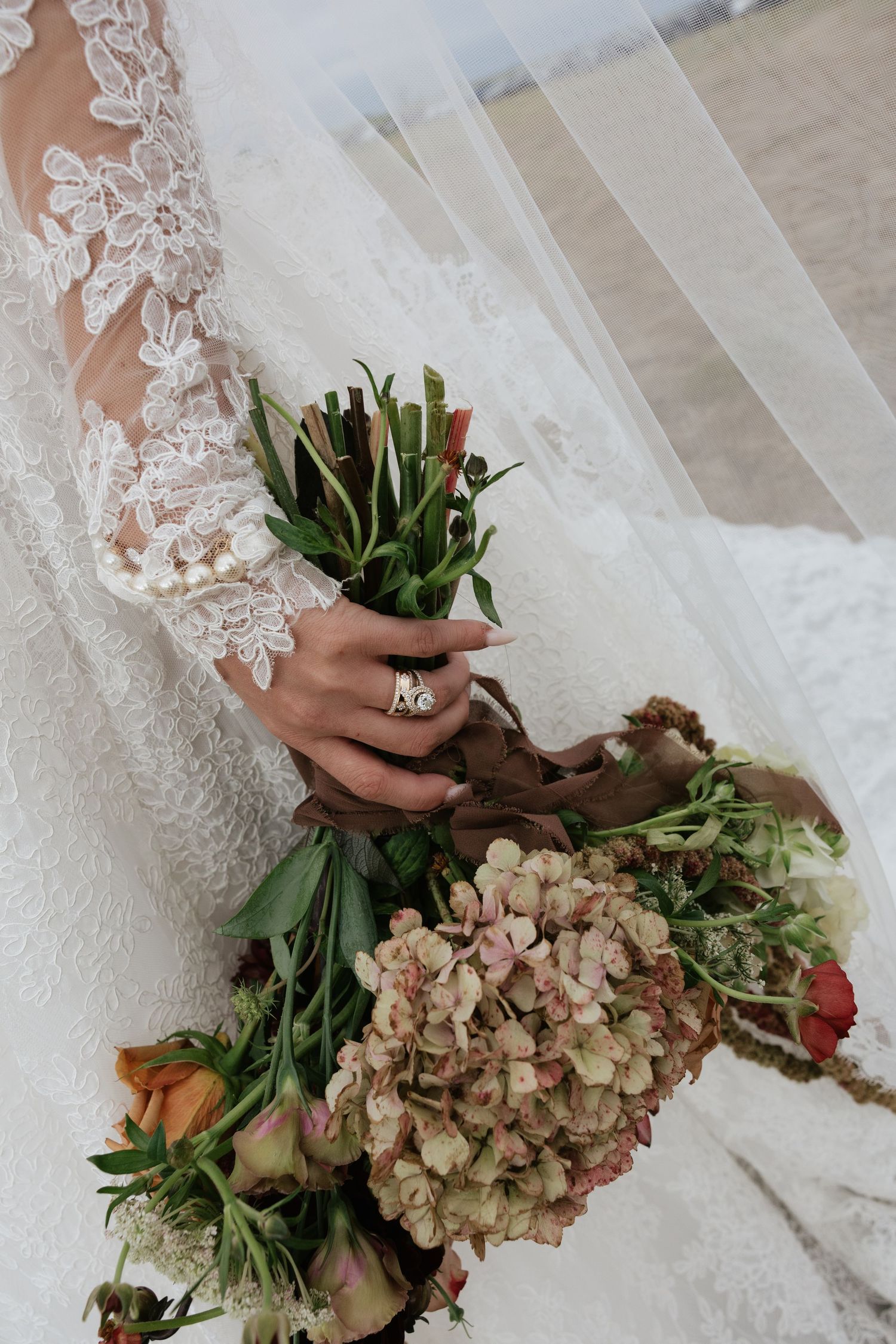 Close-up of bride in lace wedding dress holding bouquet of hydrangeas and roses with diamond ring visible.
