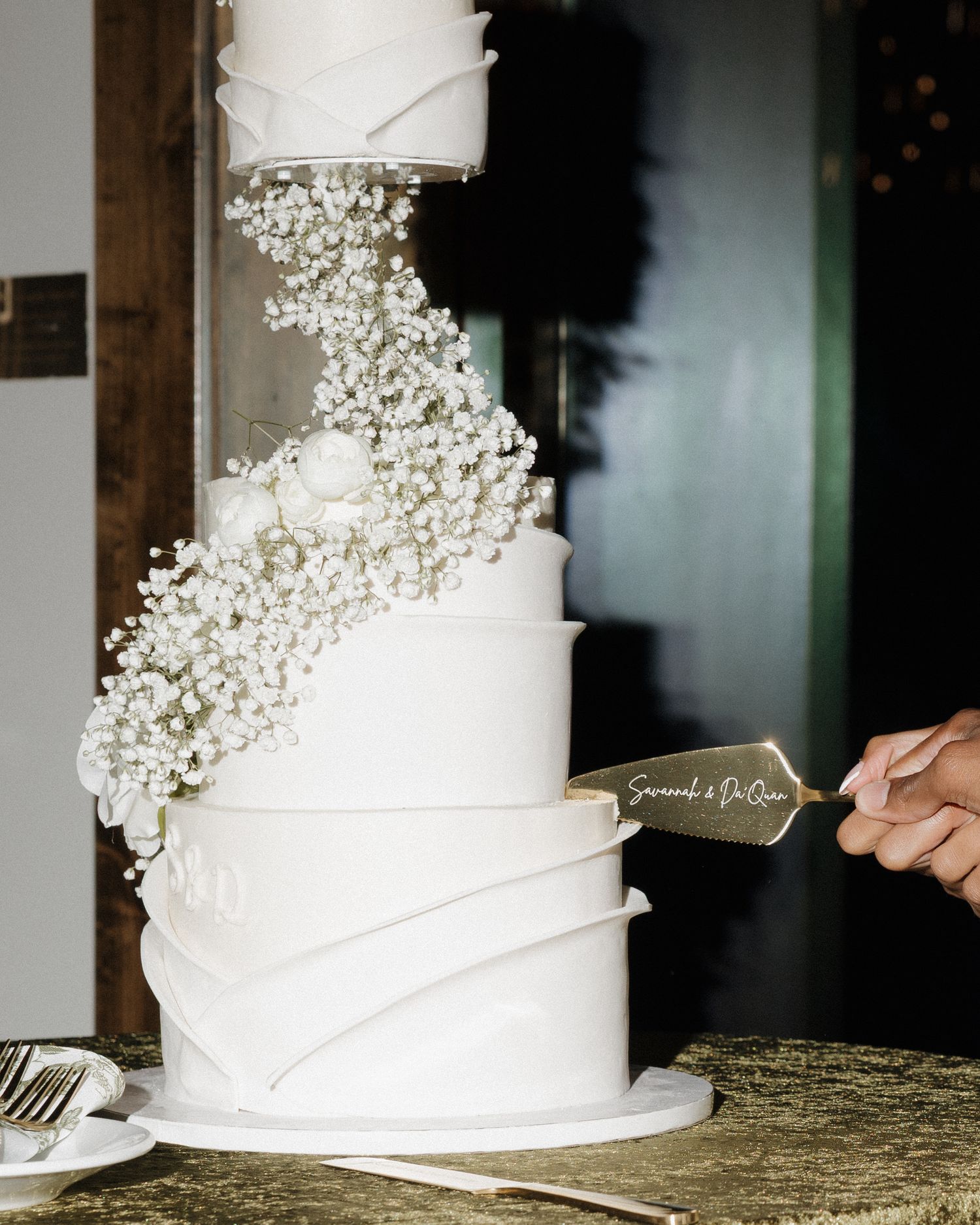 Elegant four-tier white wedding cake adorned with cascading baby's breath flowers being cut.