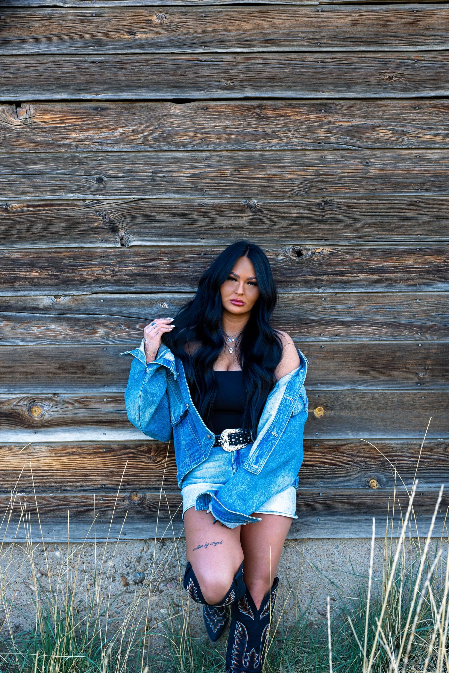 Woman in denim jacket, black bodysuit, and cowboy boots posing against rustic wooden wall.