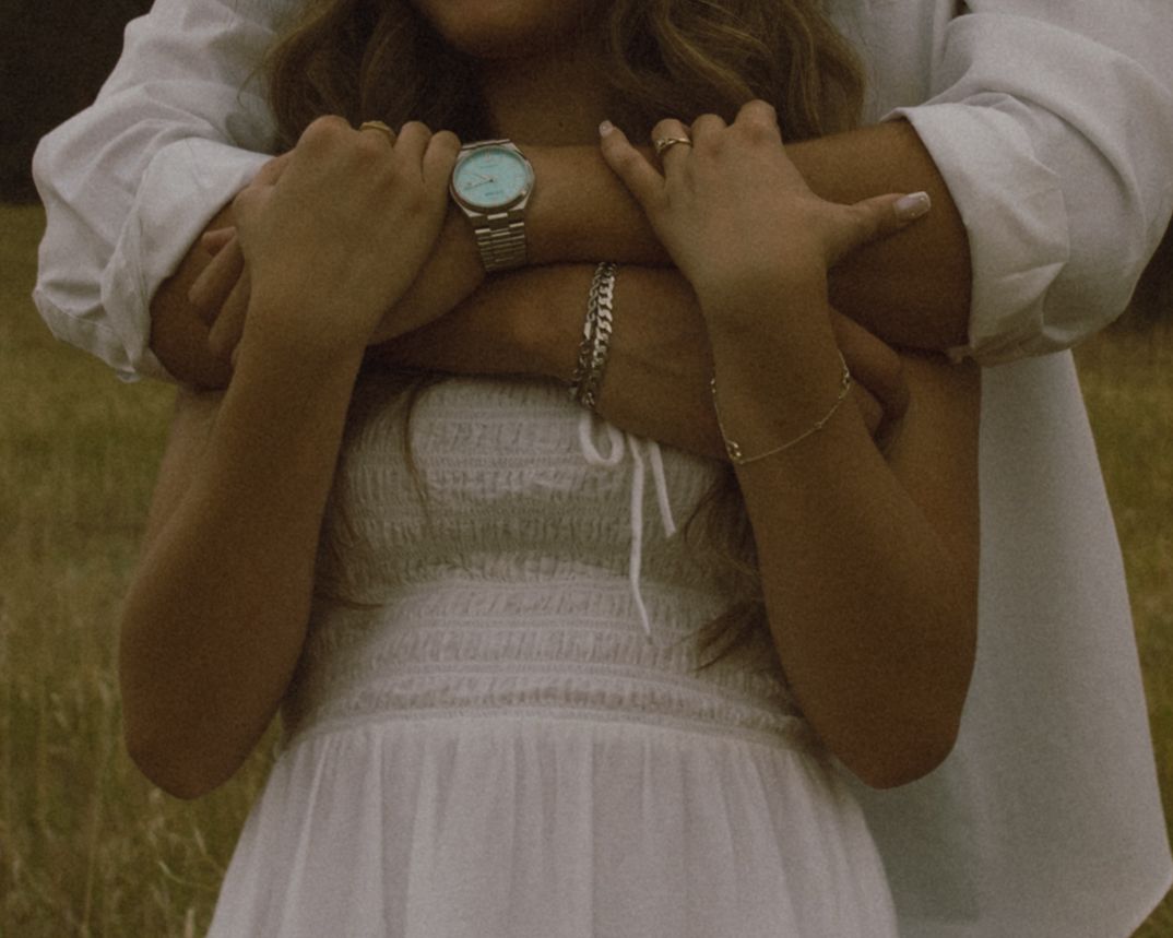 Couple embracing outdoors, woman wearing white smocked dress and teal watch, man in white shirt.