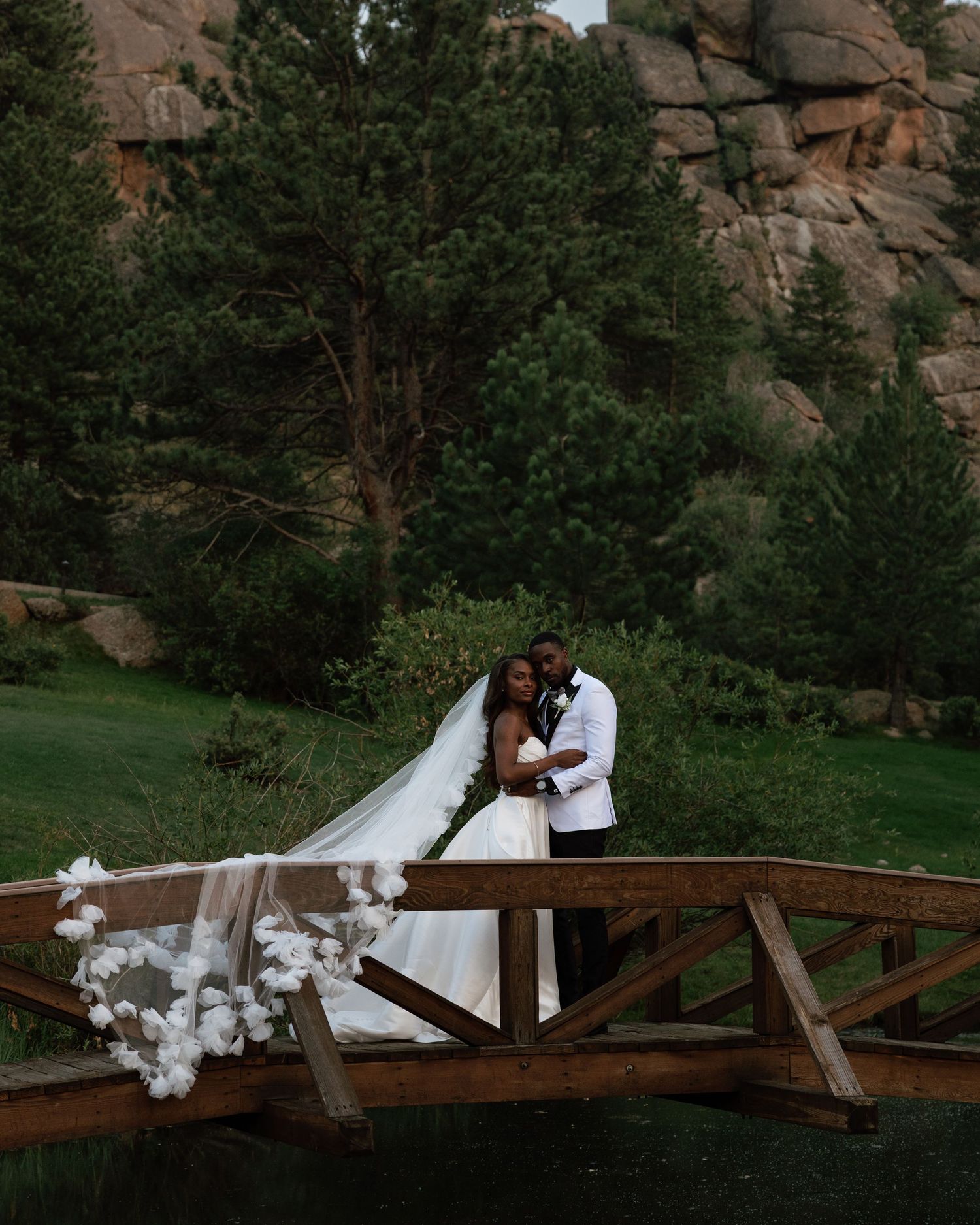 Bride and groom embrace on a wooden bridge surrounded by pine trees and rocky mountains.