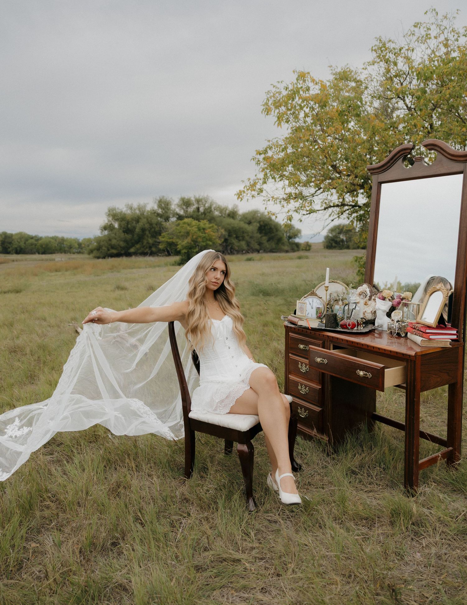 Bride in white mini wedding dress with flowing veil sits on chair beside vintage vanity in field.