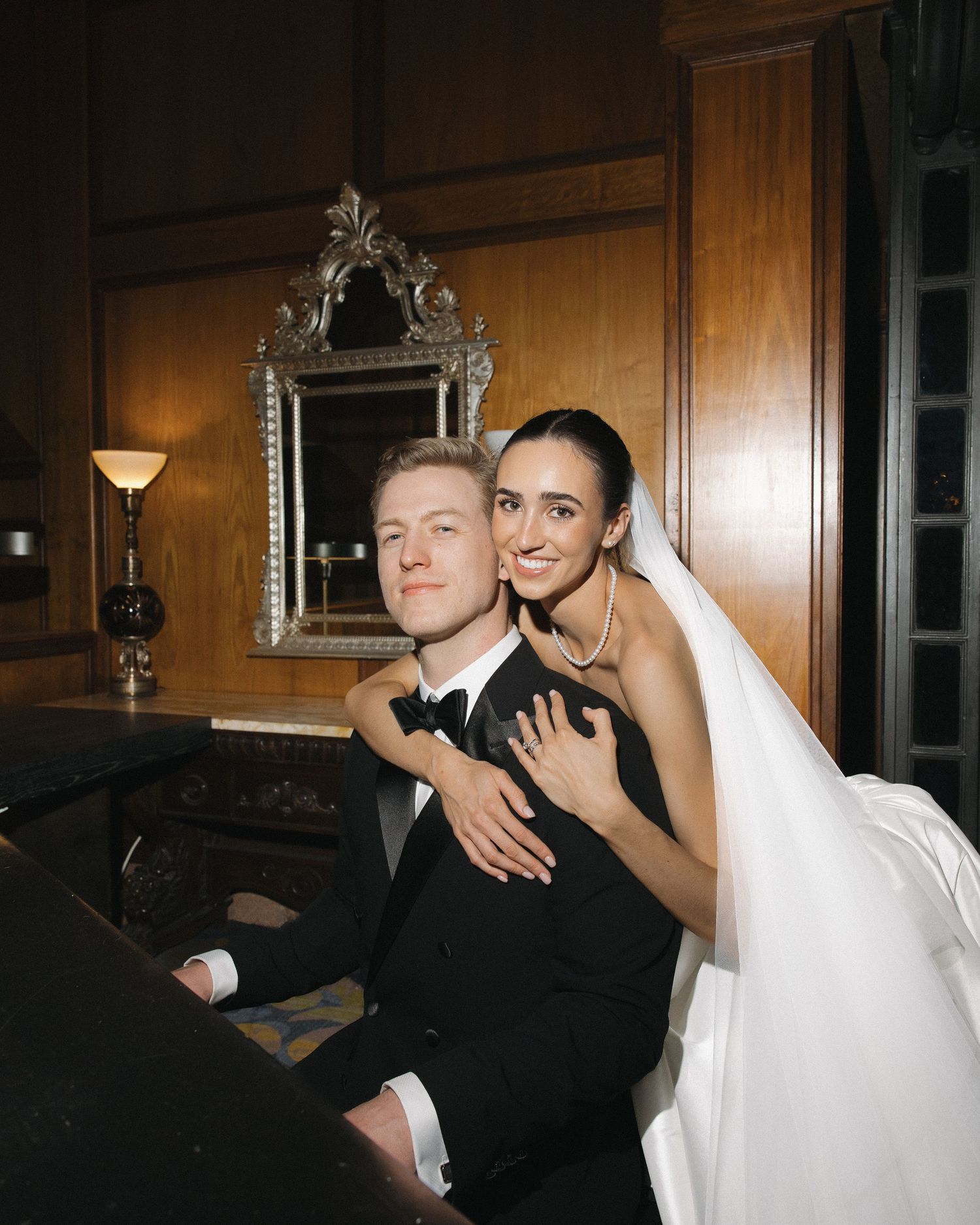 Bride and groom posing together at a grand piano in an elegant wood-paneled room.
