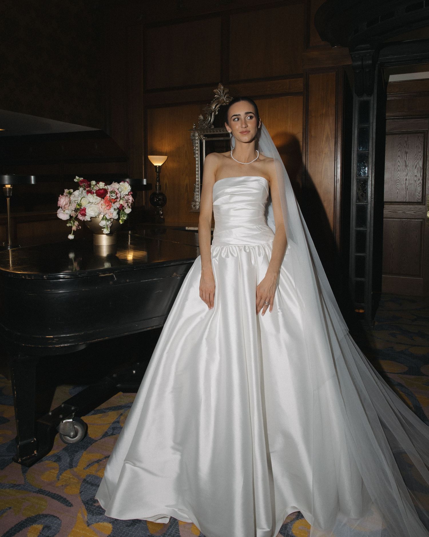 Bride in a strapless satin ballgown with cathedral veil standing beside a grand piano.