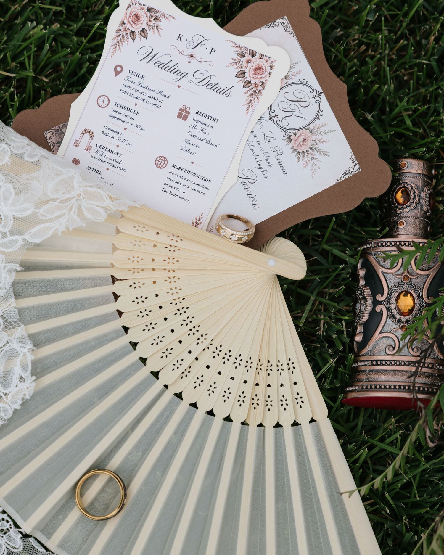 Wedding flat lay with ivory hand fan, gold rings, ornate goblet, and floral wedding details card on grass.
