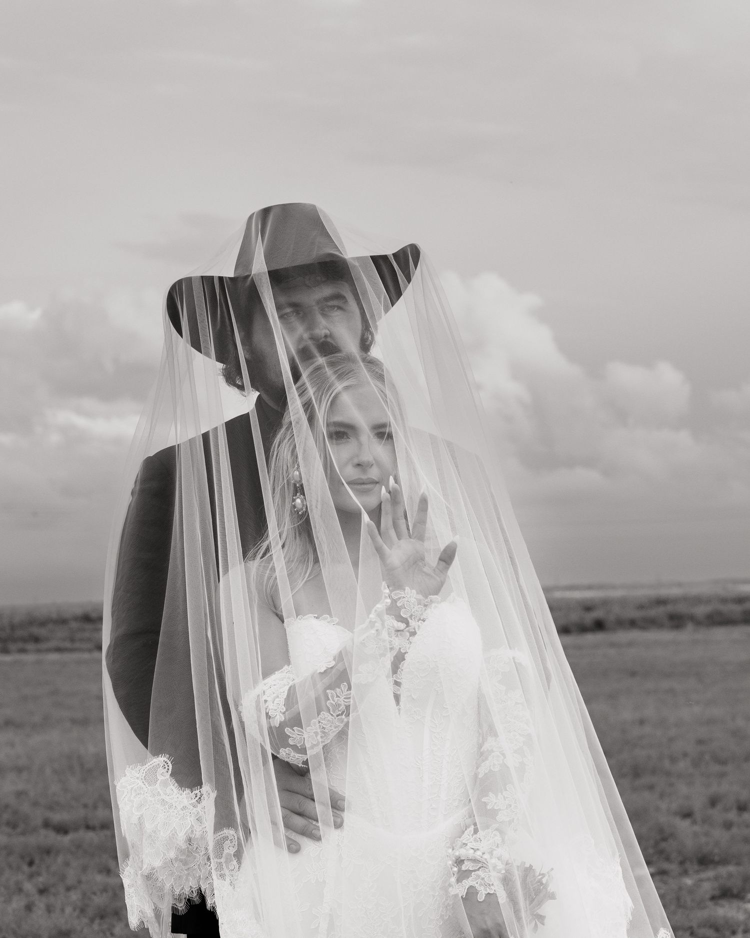 Black and white portrait of bride and groom under lace veil with dramatic cloudy sky backdrop.