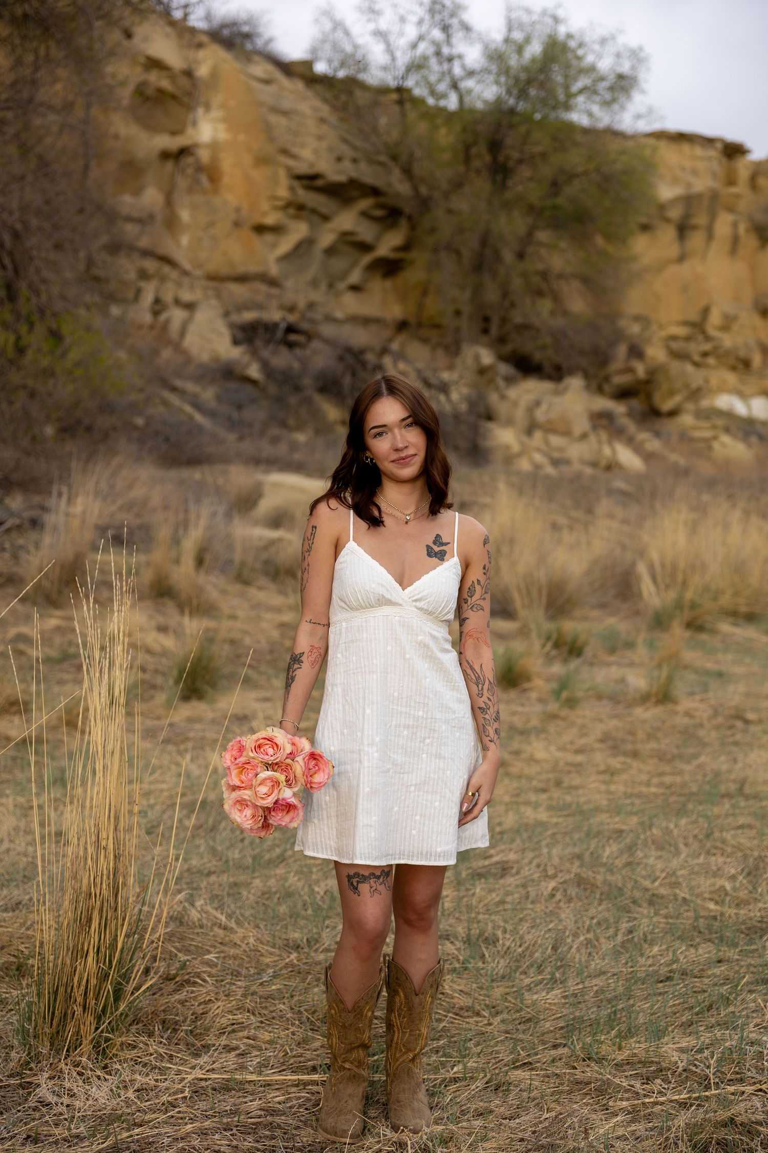 Tattooed bride in short white dress and cowboy boots holds peach bouquet in desert landscape. Engagement photography