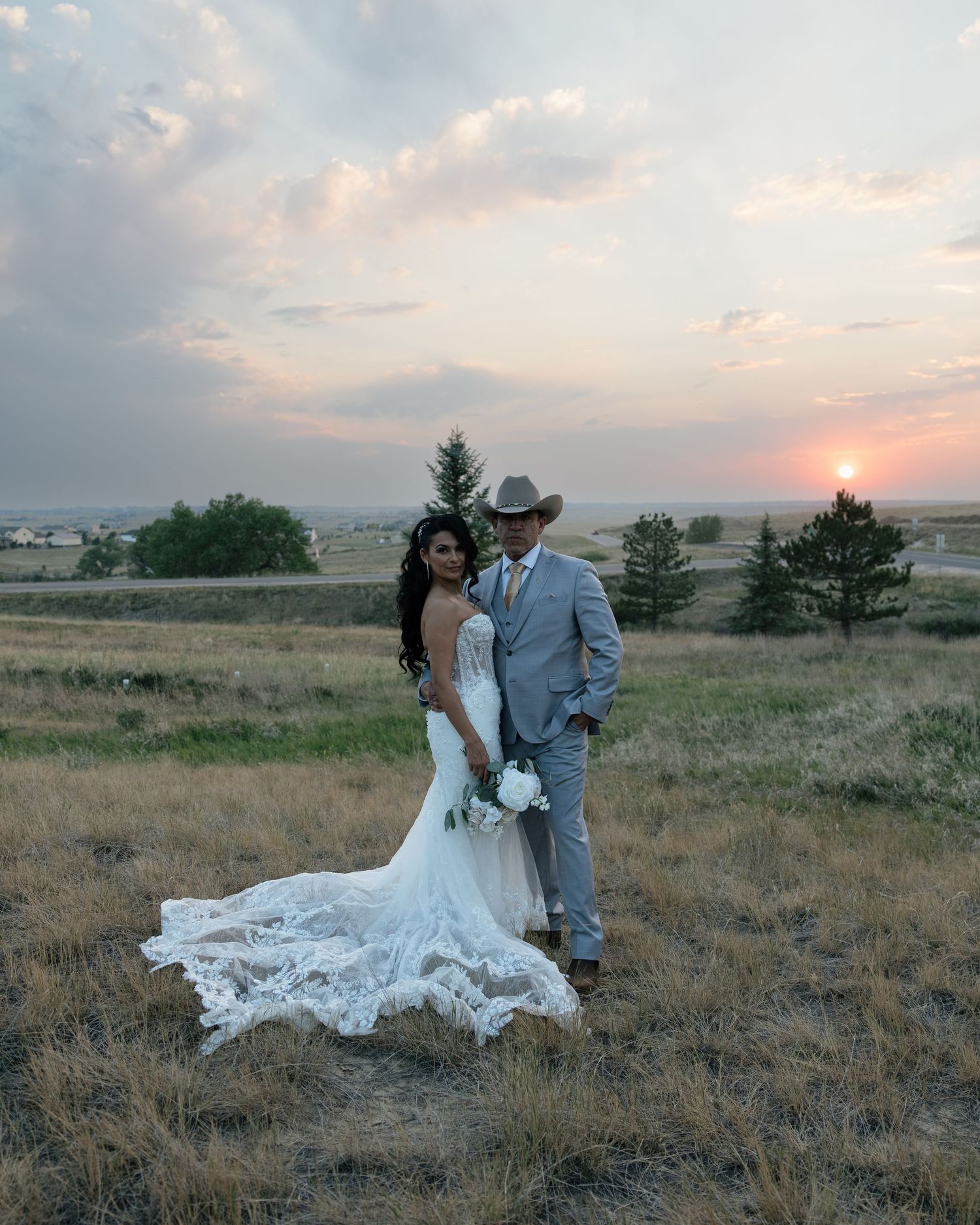 Bride in lace mermaid gown and groom in cowboy hat pose at sunset in golden field.