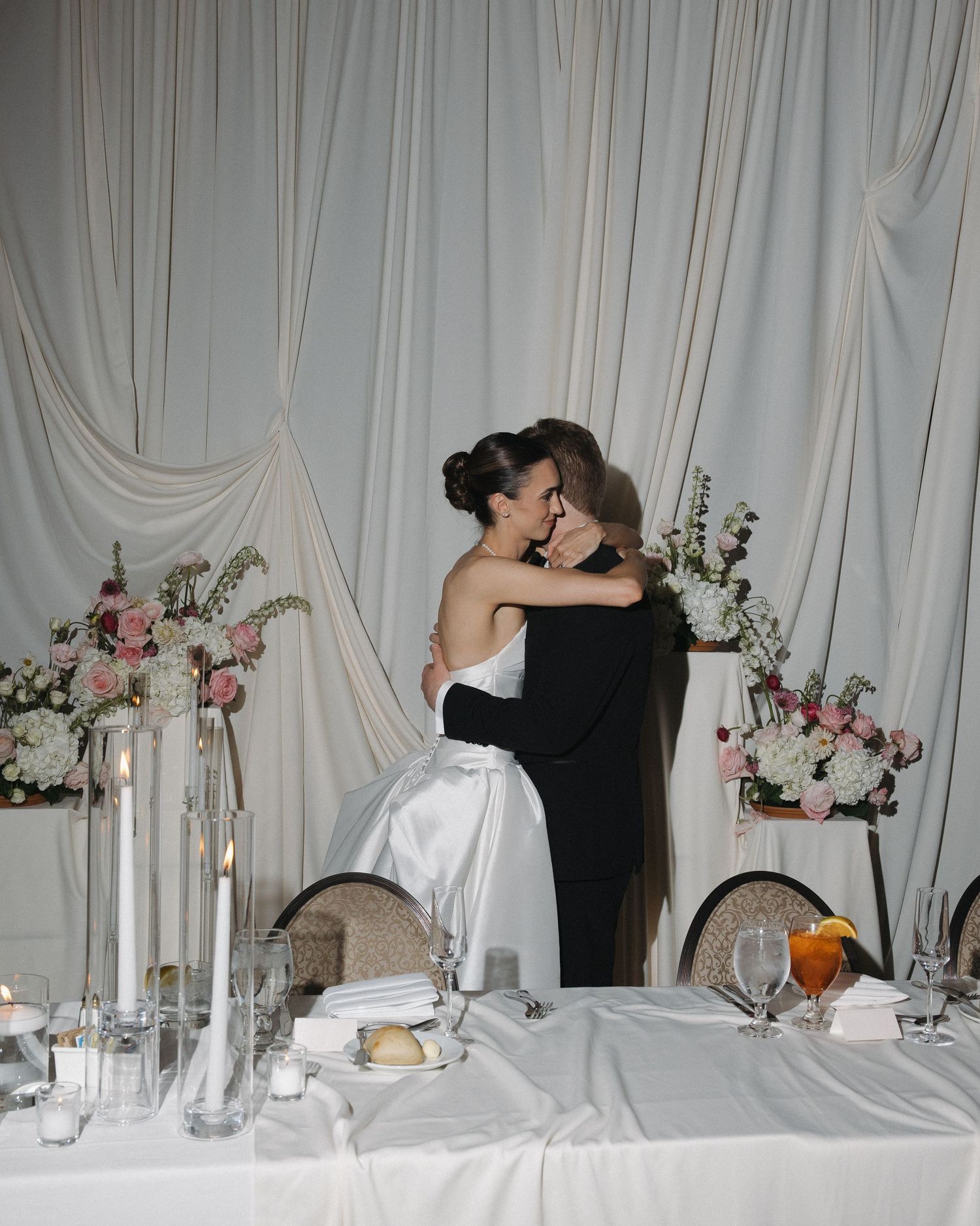 Newlyweds sharing an embrace at wedding reception table with floral arrangements and candles.