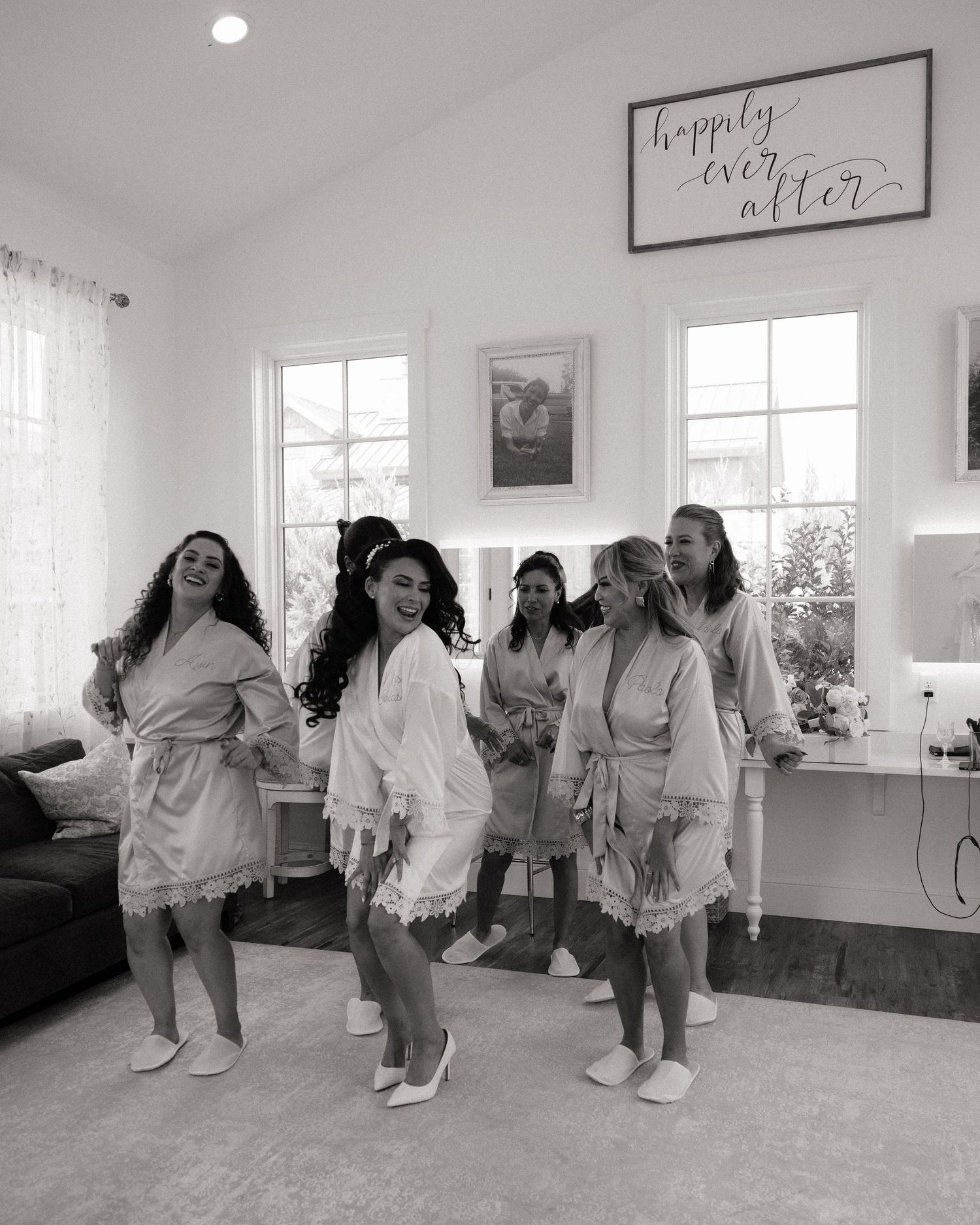 Bridesmaids in matching robes and slippers dance joyfully in bridal suite under happily ever after sign.