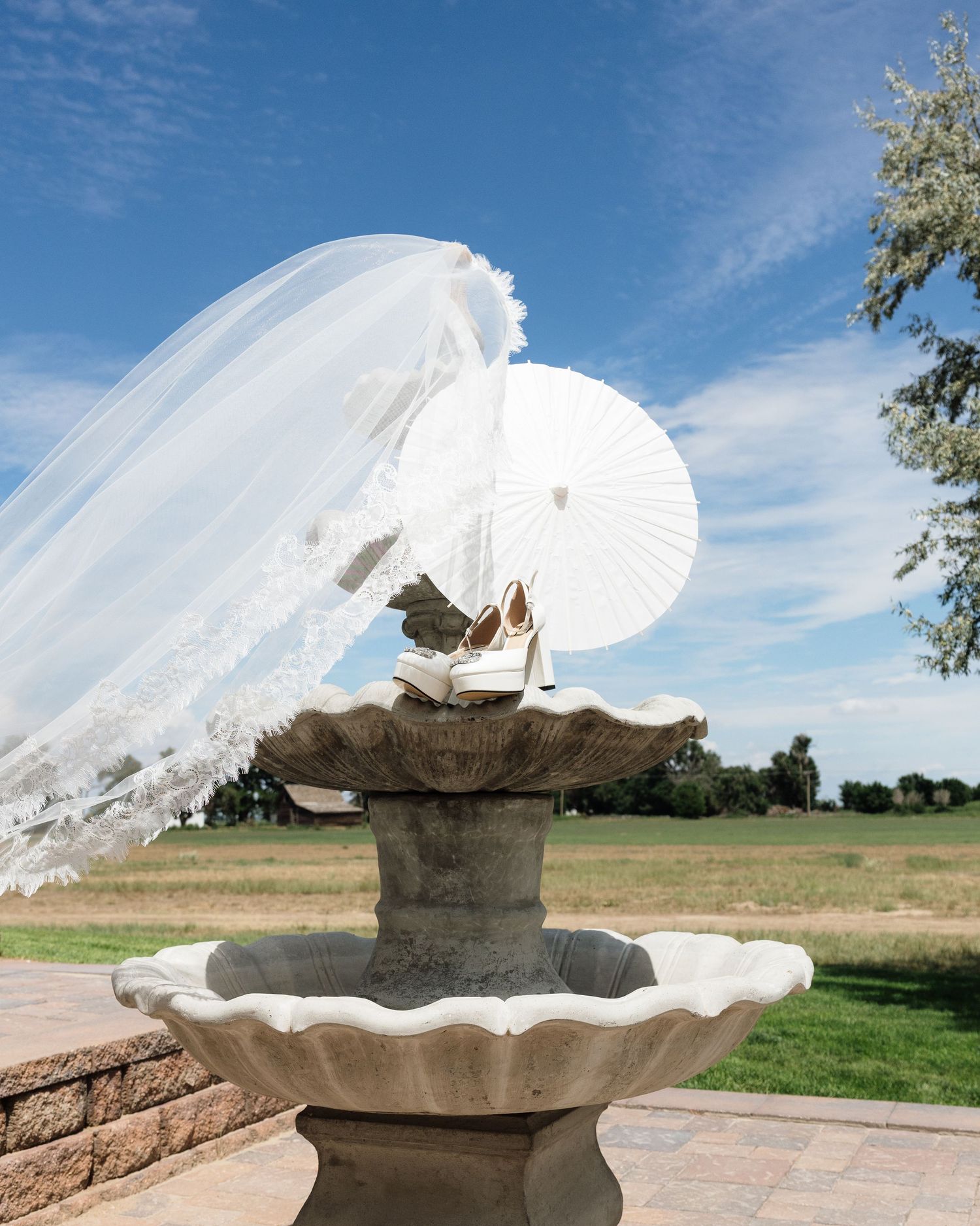Bridal veil billowing in wind over stone fountain with white parasol and wedding shoes on sunny day.